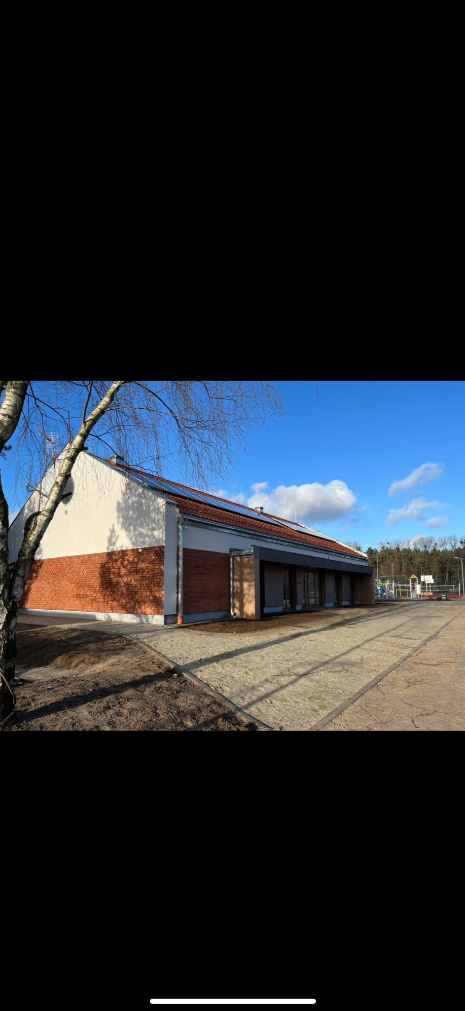 Nowoczesny budynek z ceglaną elewacją i panelami słonecznymi na dachu, otoczony terenem z placem zabaw w tle, sfotografowany w słoneczny dzień.