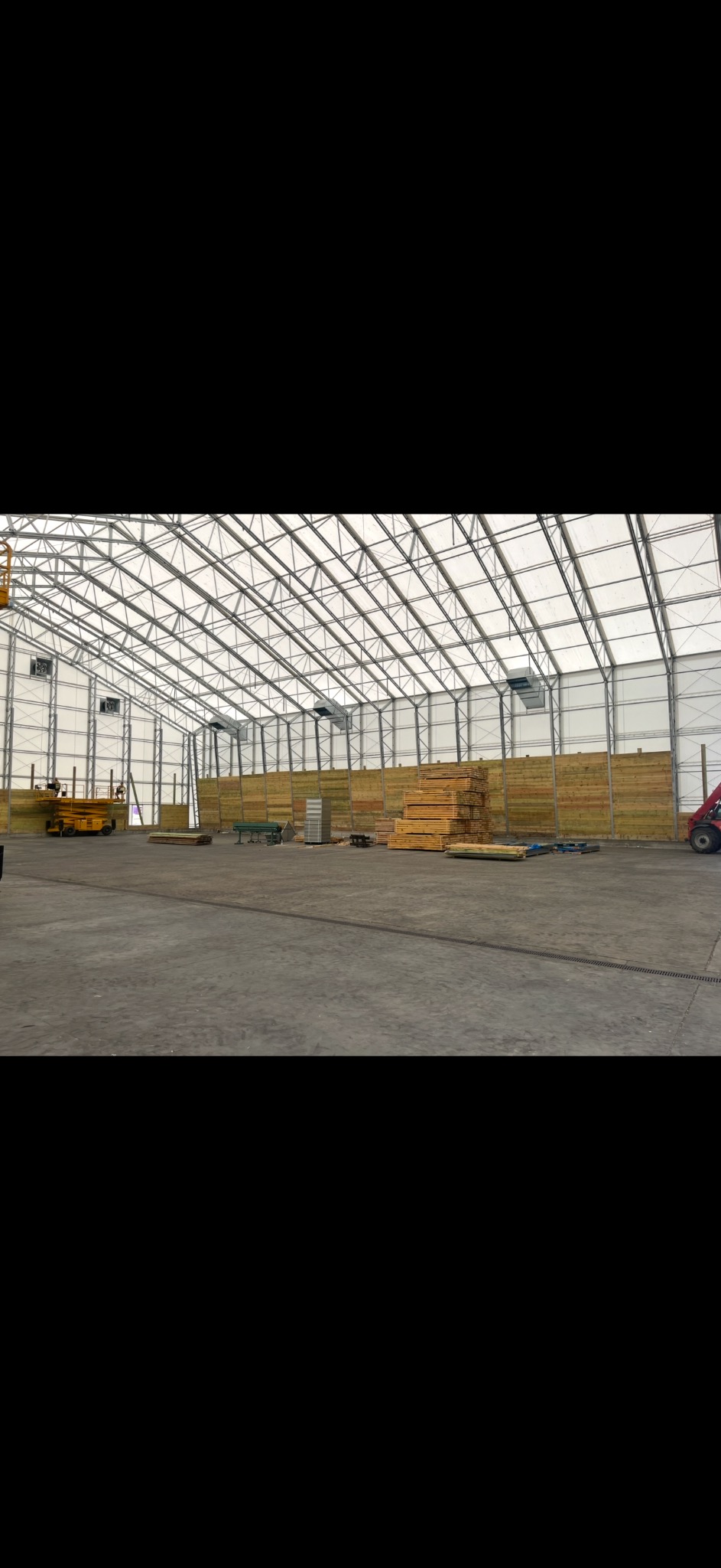 Wnętrze hali w trakcie budowy: stalowa konstrukcja, drewniane ściany, palety z drewnem, podnośnik i wózek widłowy. Widok na plac budowy wewnątrz hali.