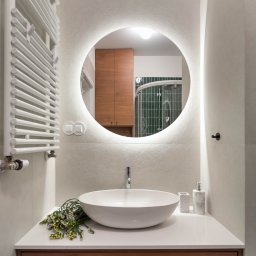 Chojecka Interior - Nowoczesna łazienka z okrągłym lustrem LED, białą umywalką nablatową i drewnianą szafką. Na blacie dekoracja z kwiatów. Widoczny grzejnik drabinkowy.