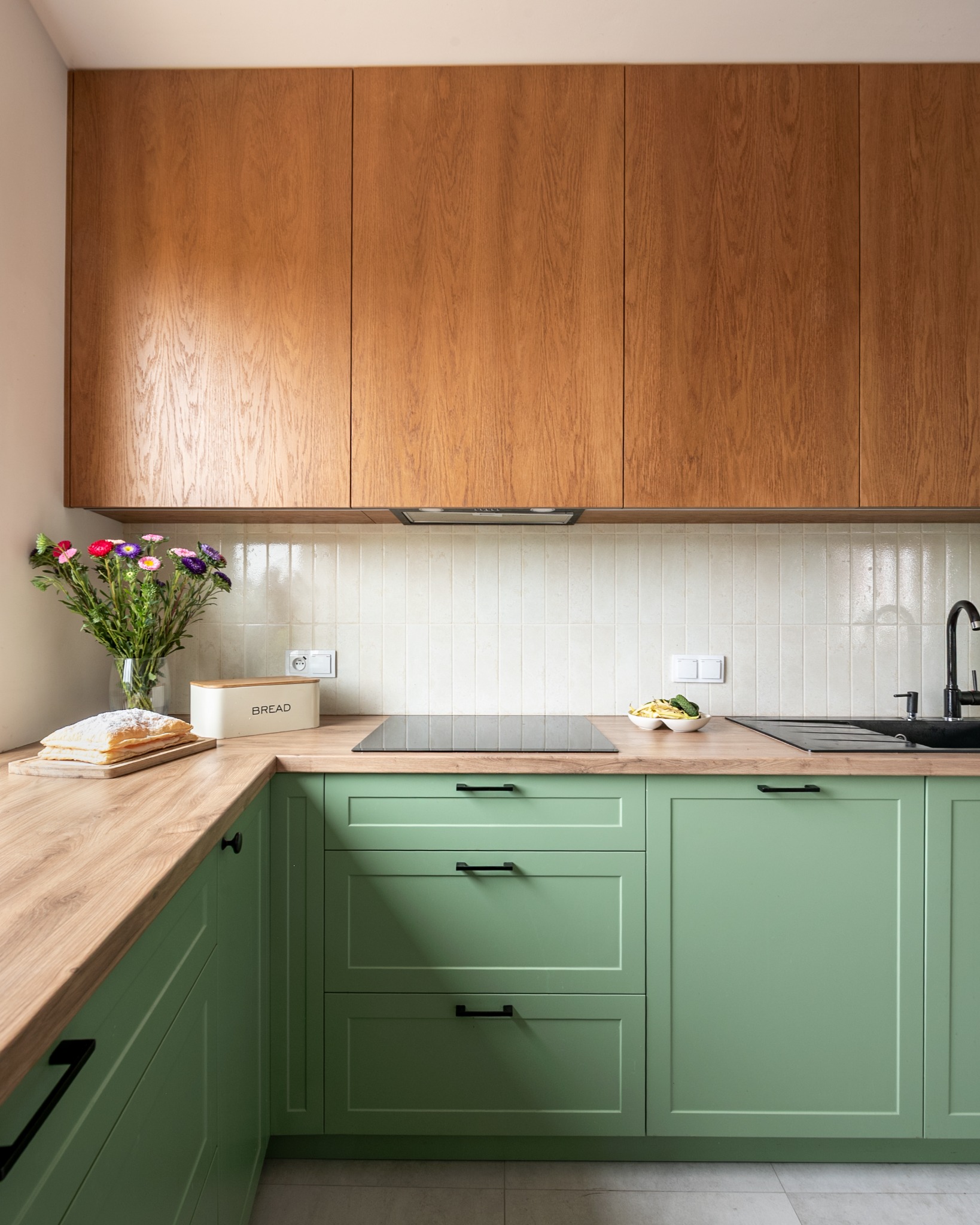 Kuchenna aranżacja z drewnianymi szafkami górnymi i zielonymi dolnymi. Na blacie kwiaty w wazonie, chlebak i deska z ciastem. Minimalistyczny design, jasne wnętrze.