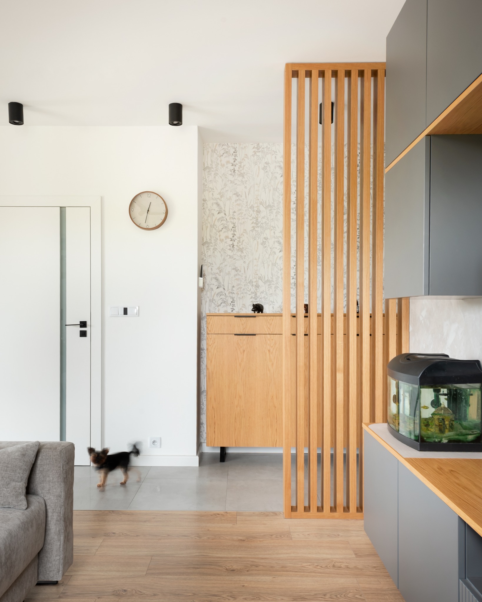 Nowoczesne wnętrze z drewnianą ażurową ścianką działową, szafką z dekoracjami i akwarium. Pies w ruchu dodaje dynamiki. Jasne kolory i minimalistyczny design.