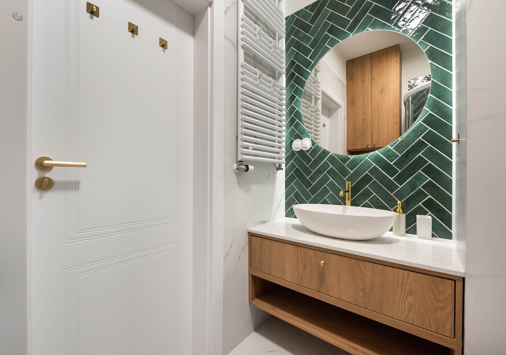 Elegancka łazienka z umywalką nablatową, drewnianą szafką, okrągłym lustrem na tle ściany z zielonej, ułożonej w jodełkę glazury. Białe drzwi i grzejnik.
