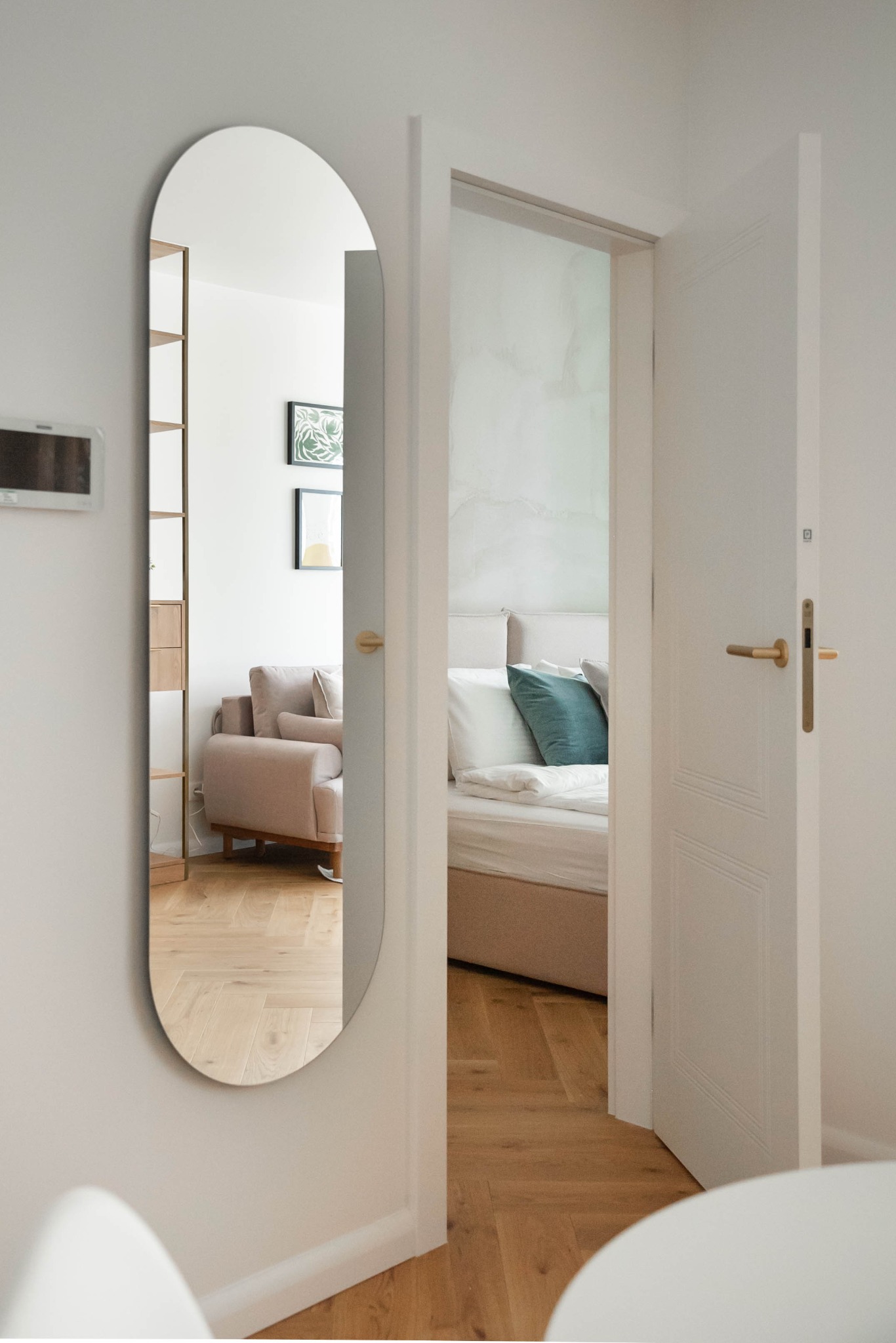 Owalne lustro na ścianie odbija pokój dzienny z beżową sofą i regałem. Widok na sypialnię przez otwarte, białe drzwi z złotą klamką. Parkiet na podłodze.
