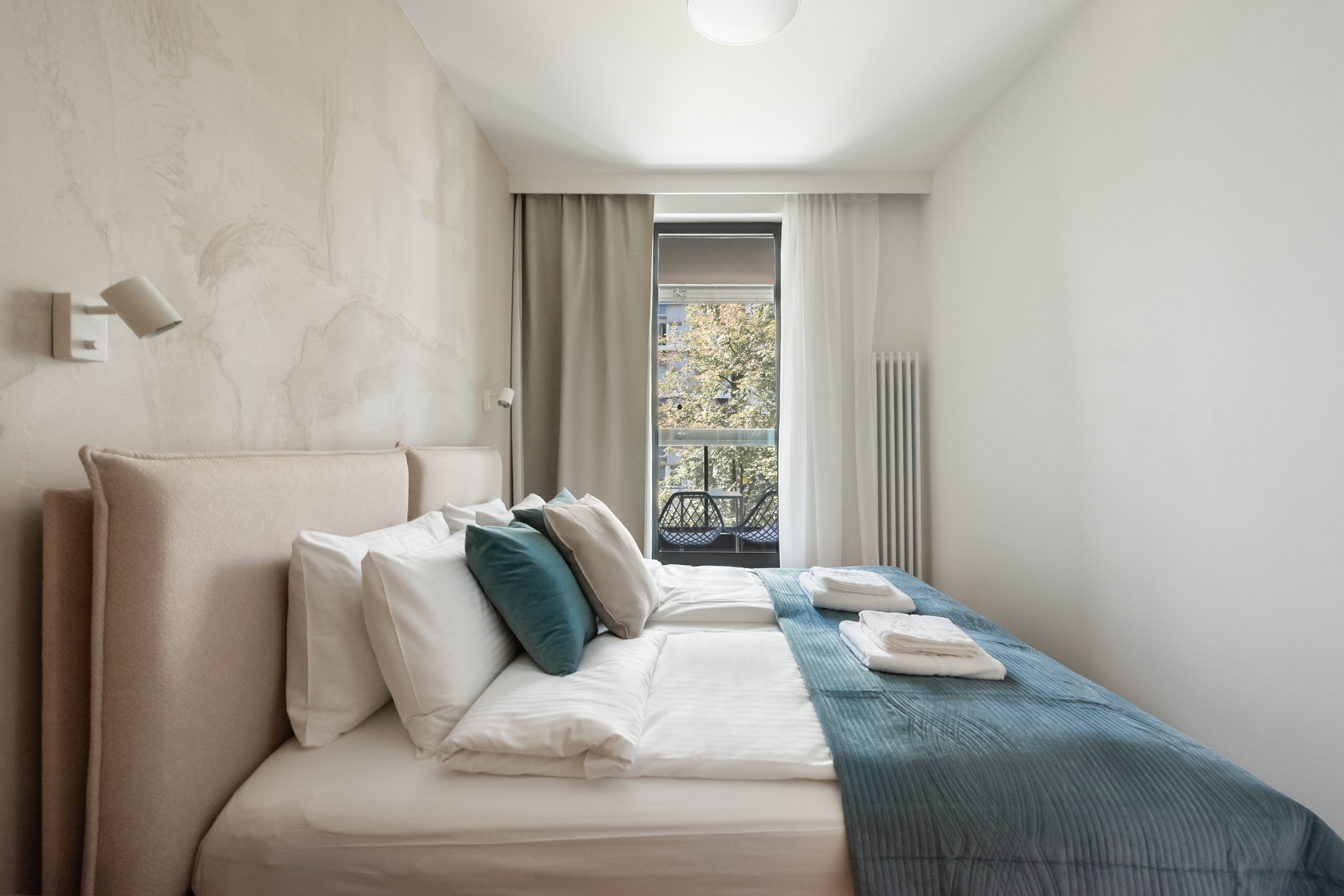 Jasna sypialnia z łóżkiem, dekoracyjną ścianą i balkonem z krzesłami. Minimalistyczny design, tekstylia w odcieniach beżu i błękitu. Widok na zieleń za oknem.