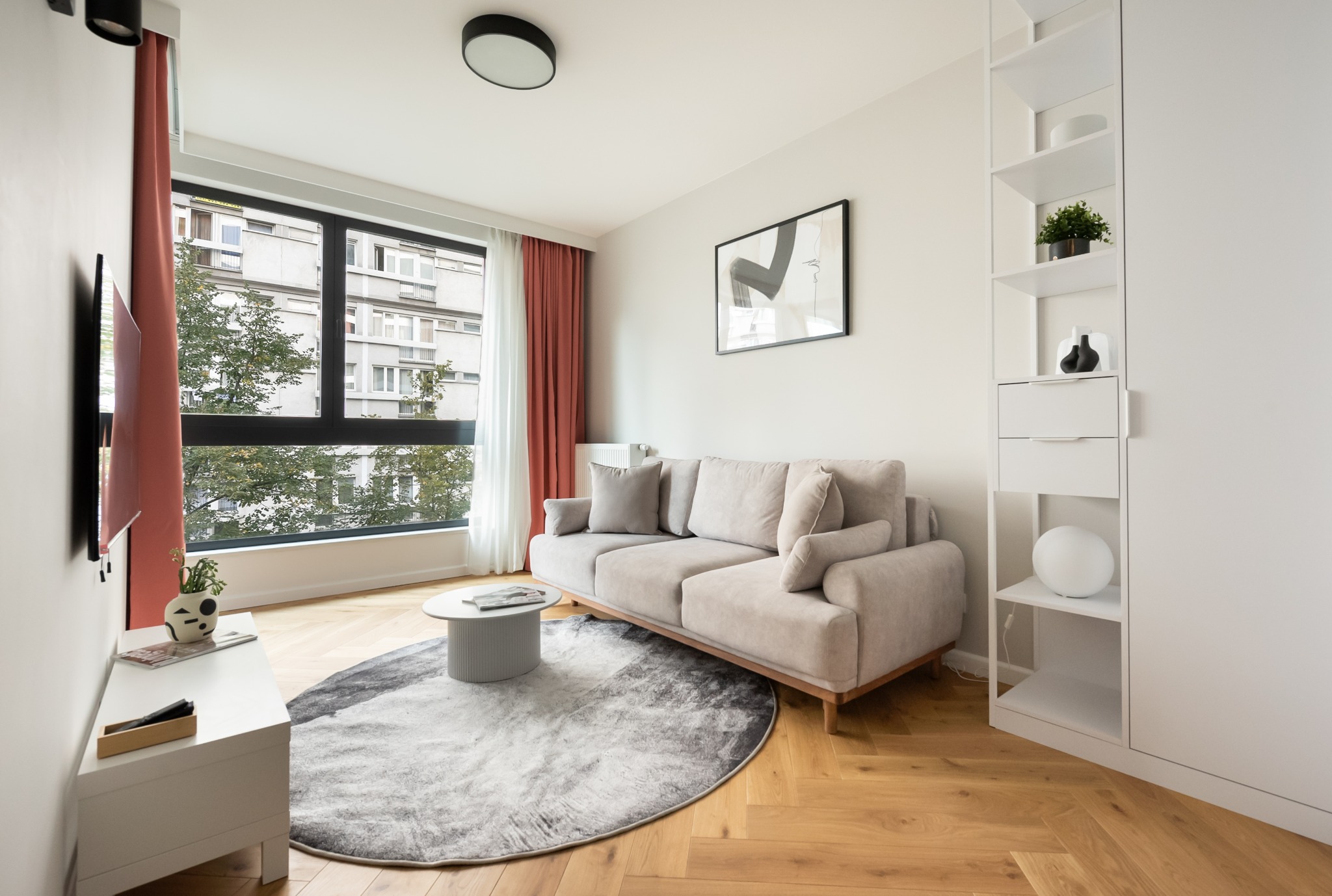 Jasny salon z beżową sofą, okrągłym dywanem i białymi meblami. Widok na budynek zza okna. Minimalistyczny wystrój, drewniana podłoga w jodełkę.