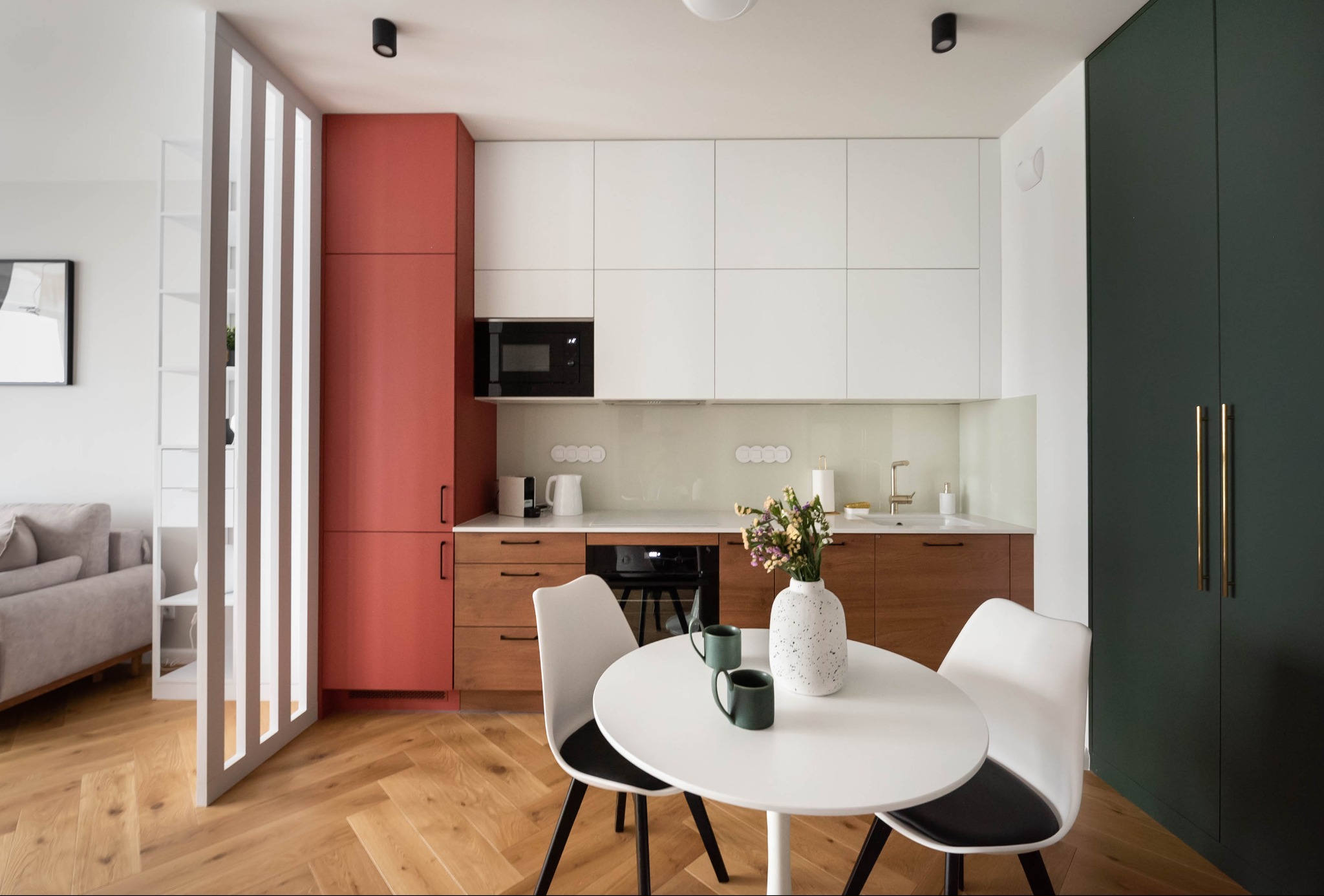 Nowoczesna kuchnia z drewnianą podłogą w jodełkę, białą zabudową i akcentami w kolorze ceglastym oraz butelkowej zieleni. Okrągły stół z kwiatami.