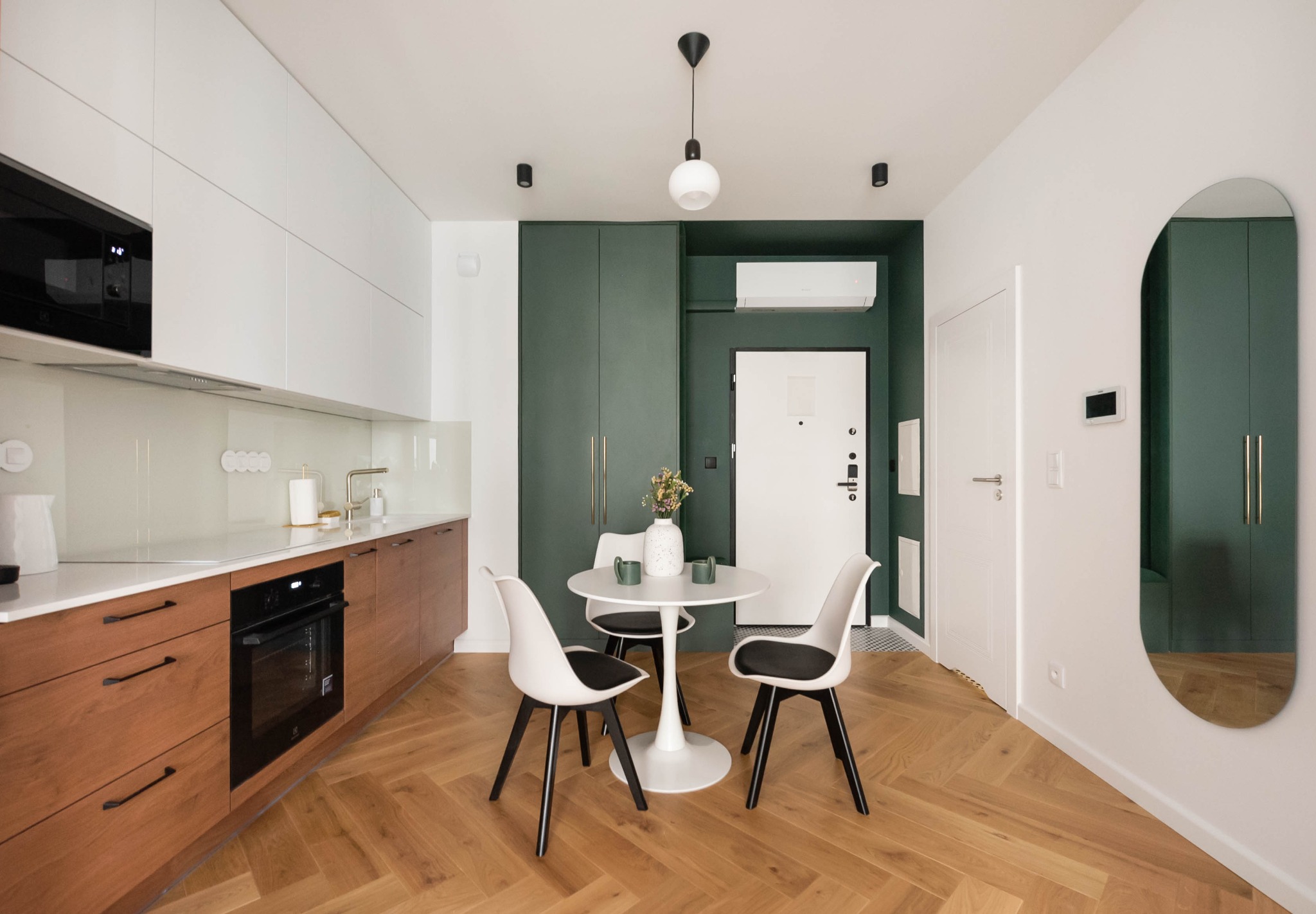Nowoczesne wnętrze: drewniana podłoga w jodełkę, białe i drewniane szafki kuchenne, okrągły stół z krzesłami, zielona ściana i szafa w zabudowie, lustro.