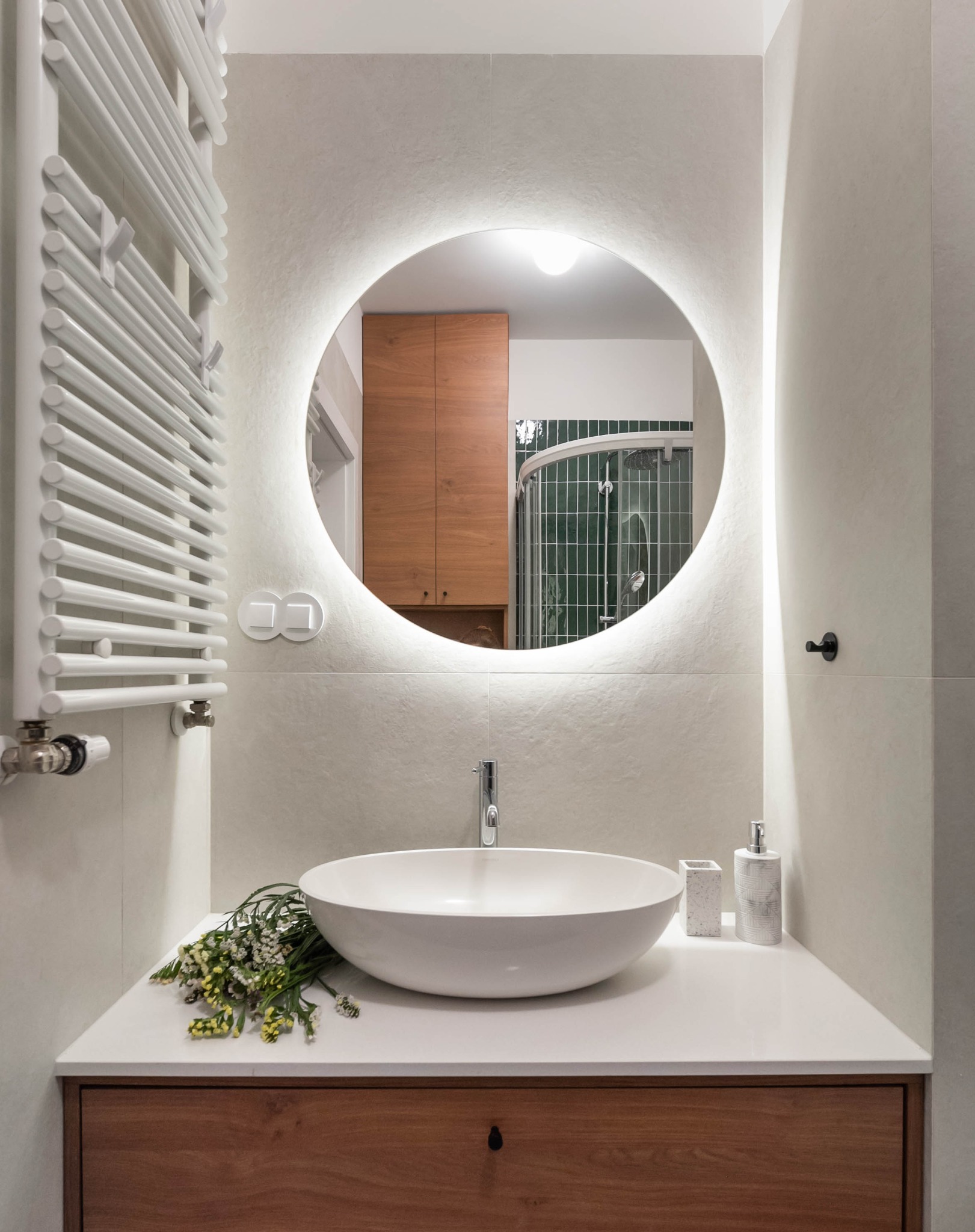 Nowoczesna łazienka z okrągłym lustrem LED, białą umywalką nablatową i drewnianą szafką. Na blacie dekoracja z kwiatów. Widoczny grzejnik drabinkowy.