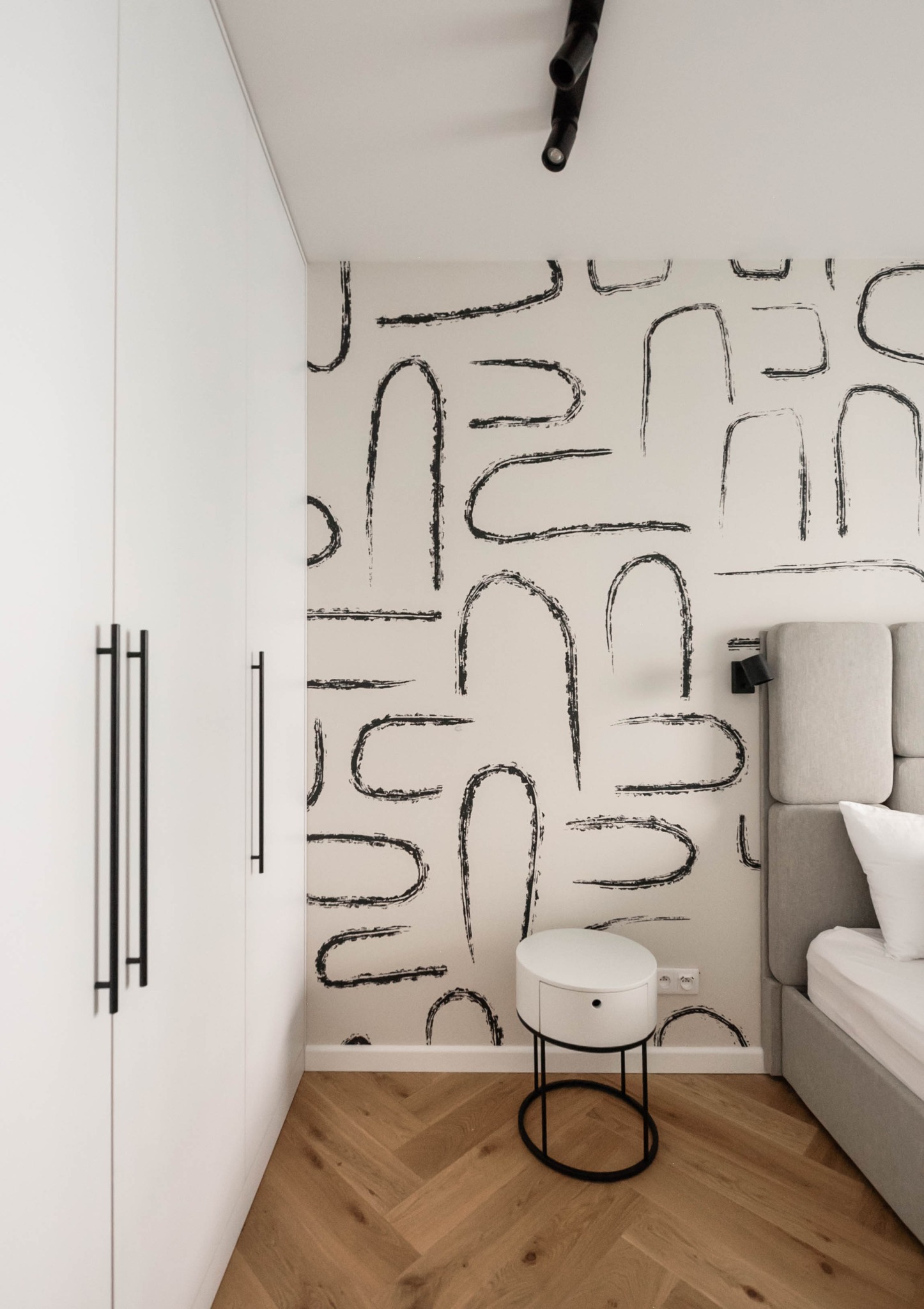 Minimalistyczna sypialnia: biała szafa z czarnymi uchwytami, ściana z abstrakcyjnym wzorem, okrągły stolik nocny i fragment łóżka z szarym zagłówkiem.