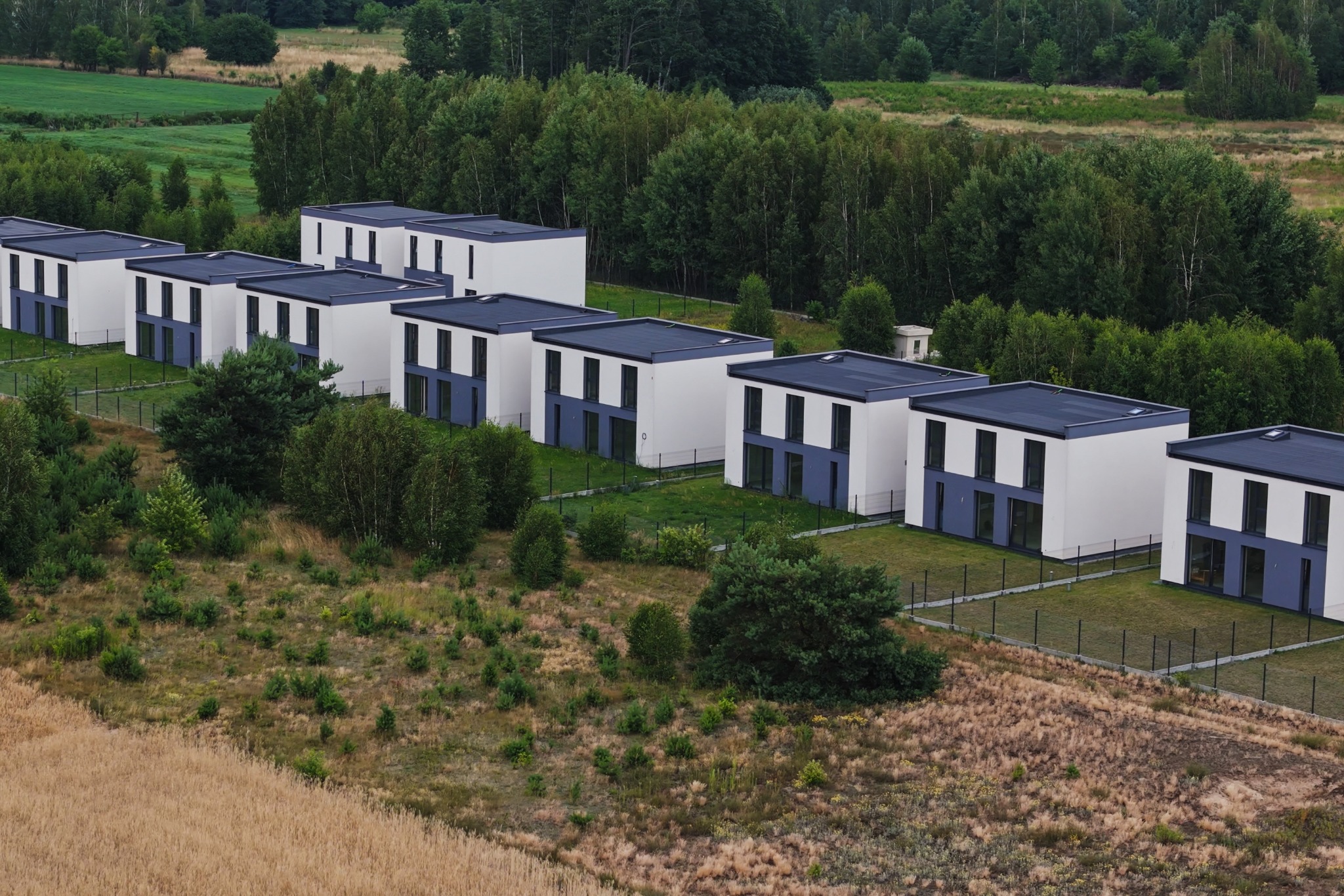 Rząd nowoczesnych, białych domów z płaskimi dachami i szarymi akcentami, otoczonych zielenią i niskim ogrodzeniem, widzianych z góry.