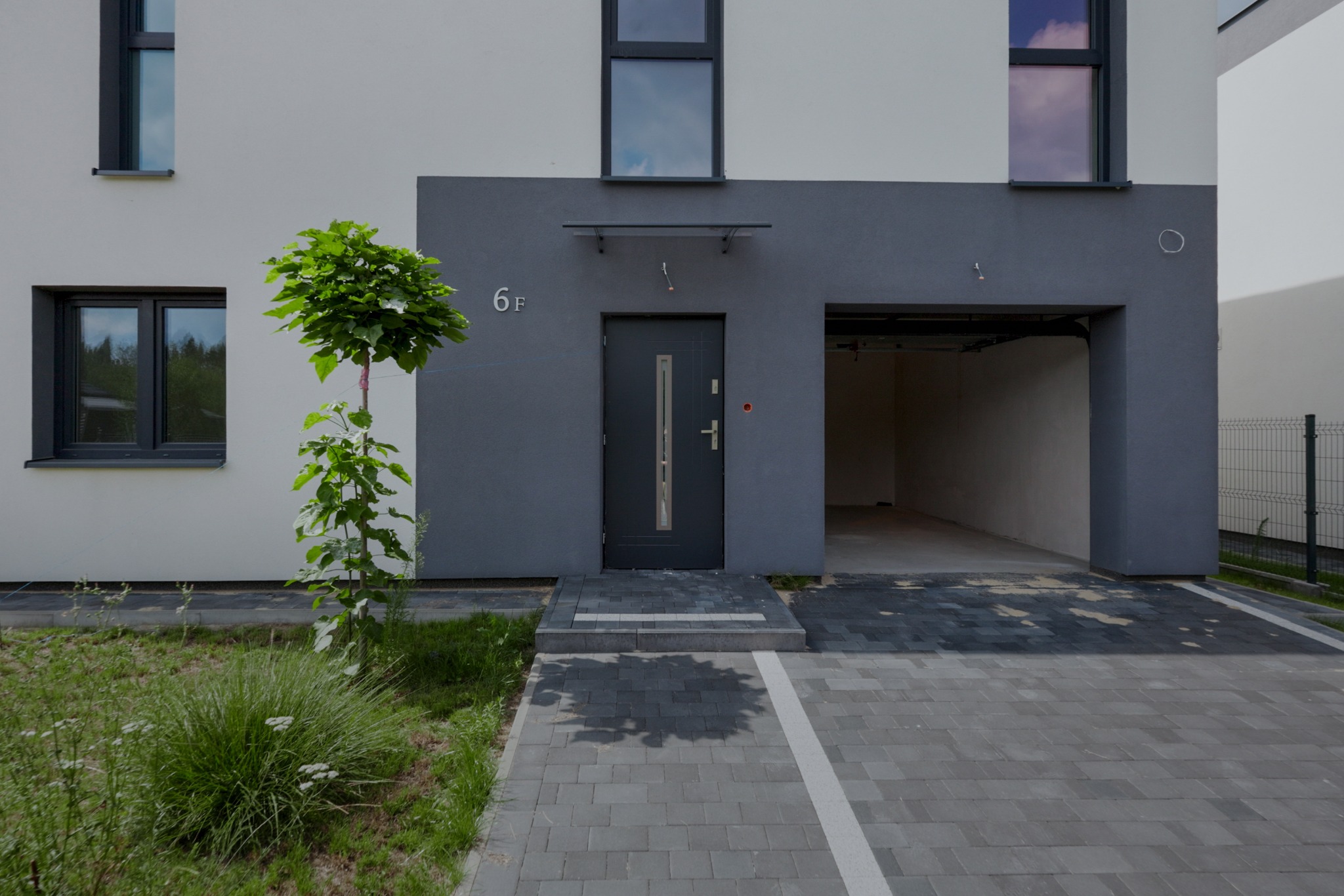 Nowoczesny budynek z szarą elewacją i numerem 6F, z wejściem i wjazdem do garażu. Kostka brukowa na podjeździe i trawnik z roślinnością.