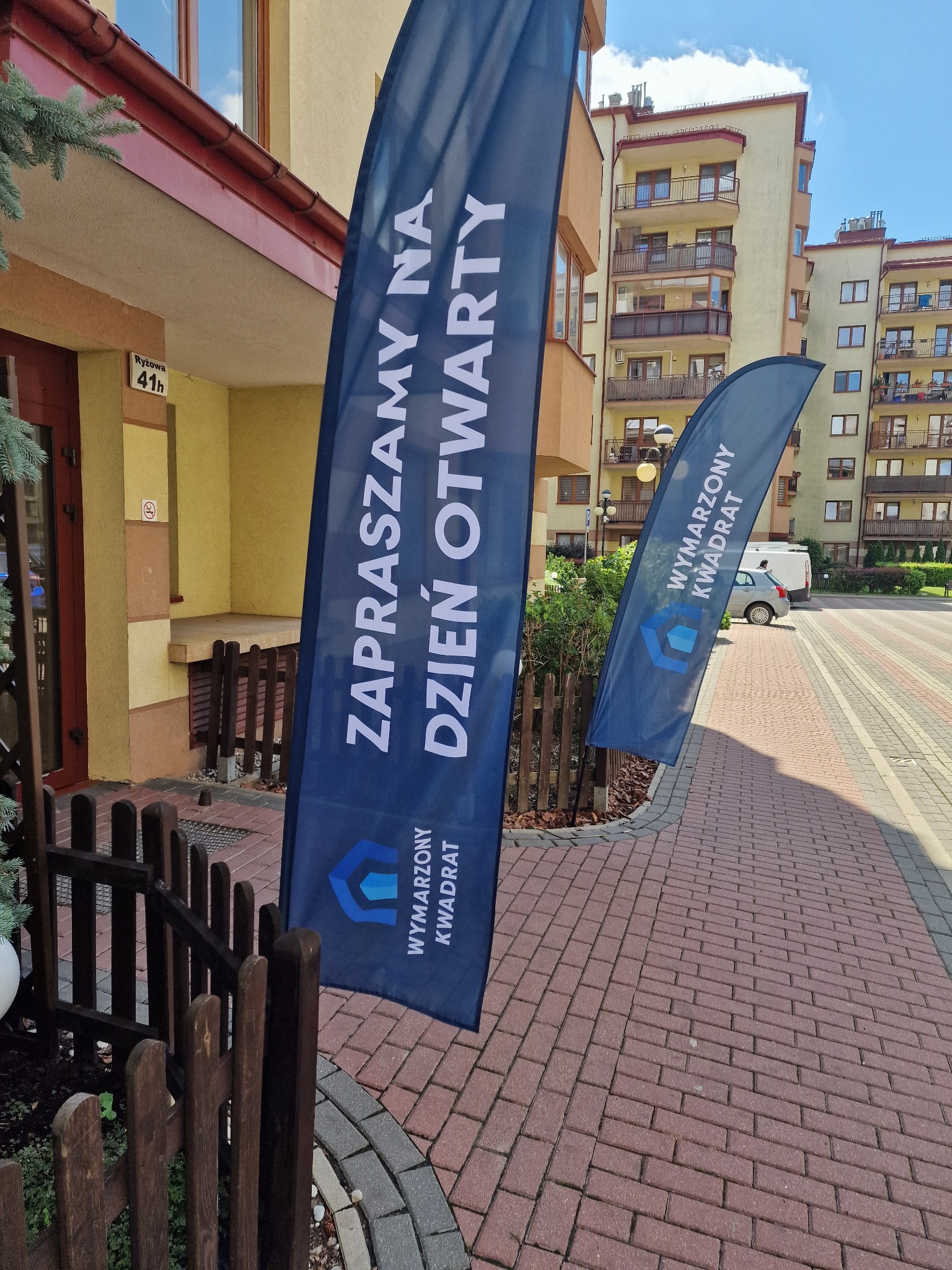 Dwa banery typu windery przed budynkiem mieszkalnym, zapraszające na dzień otwarty. Widoczny adres Ryżowa 41h.