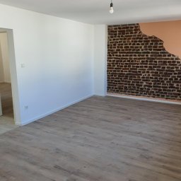 Firmanty Construction & Design - Pokój po remoncie: panele podłogowe, białe ściany i fragment ściany z cegły z odsłoniętym tynkiem w kolorze brzoskwiniowym. Widoczna żarówka na kablu.