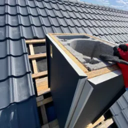 Montaż obróbki komina na dachu krytym szarą dachówką. Widoczna więźba i ocieplenie. Praca dekarza w trakcie, rękawice ochronne. Słoneczny dzień, błękitne niebo.