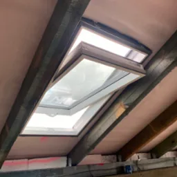 Otwarty, biały okno dachowe zamontowane w skośnym dachu z widoczną konstrukcją drewnianą i izolacją. Widok od wewnątrz, w tle jasne niebo.