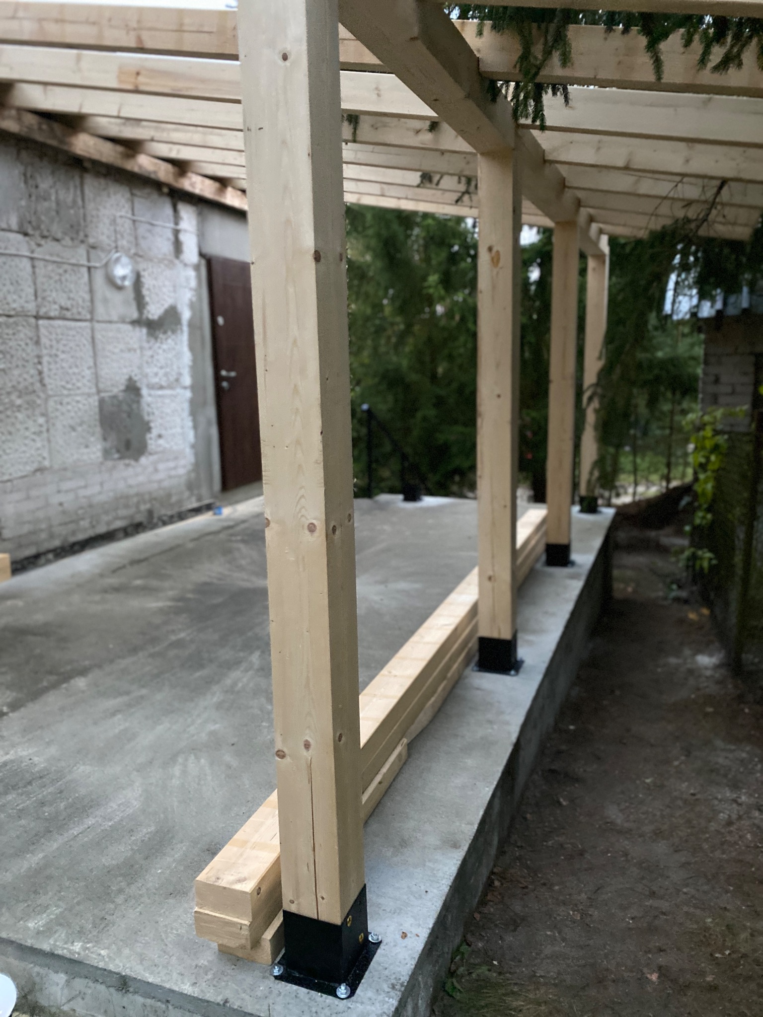 Drewniana konstrukcja z belek, wsparta na metalowych podstawach, w trakcie montażu. Widoczne elementy dachu i betonowa podstawa.