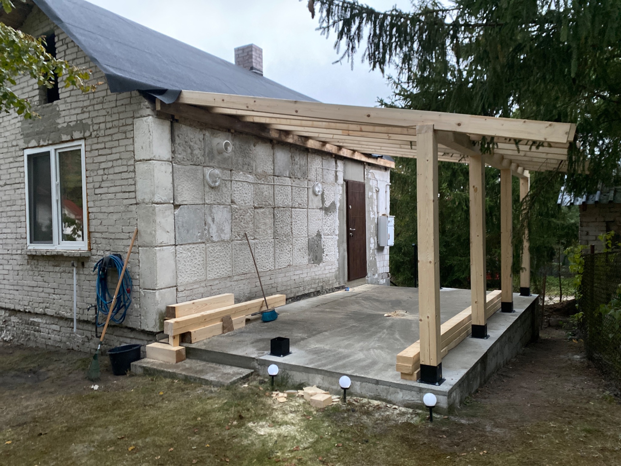 Budowa drewnianej konstrukcji zadaszenia przy murowanym budynku z oknem i dachem pokrytym papą. Widoczne narzędzia i materiały budowlane.