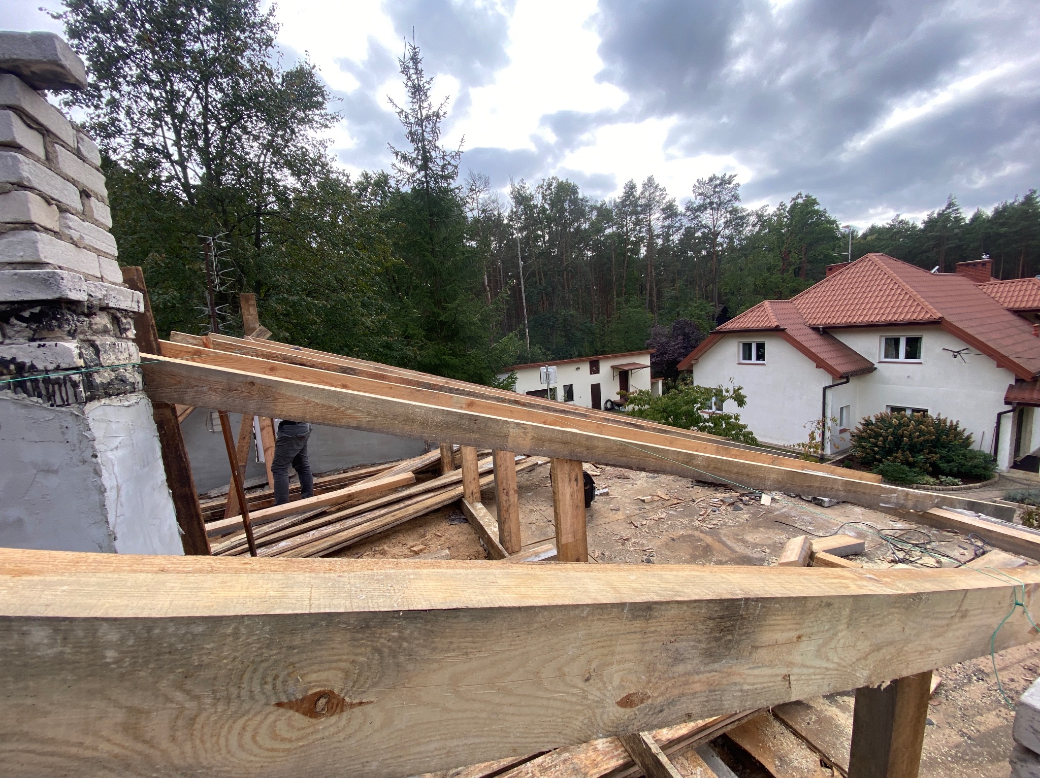 Montaż drewnianej konstrukcji dachu domu jednorodzinnego. Widoczny fragment komina i elementy więźby na tle zieleni drzew i pochmurnego nieba.