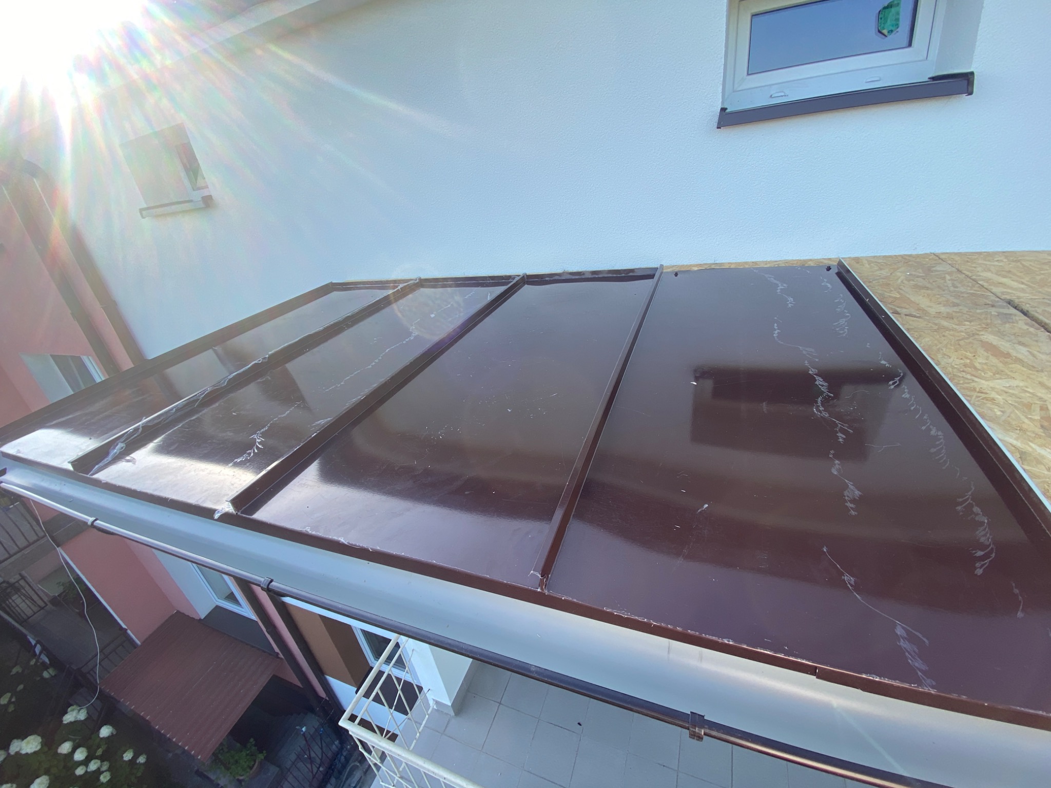 Błyszczący, brązowy dach z blachy trapezowej w trakcie montażu, widok z góry. Częściowo widoczne rynny i fragment elewacji z oknem. Słoneczne oświetlenie.