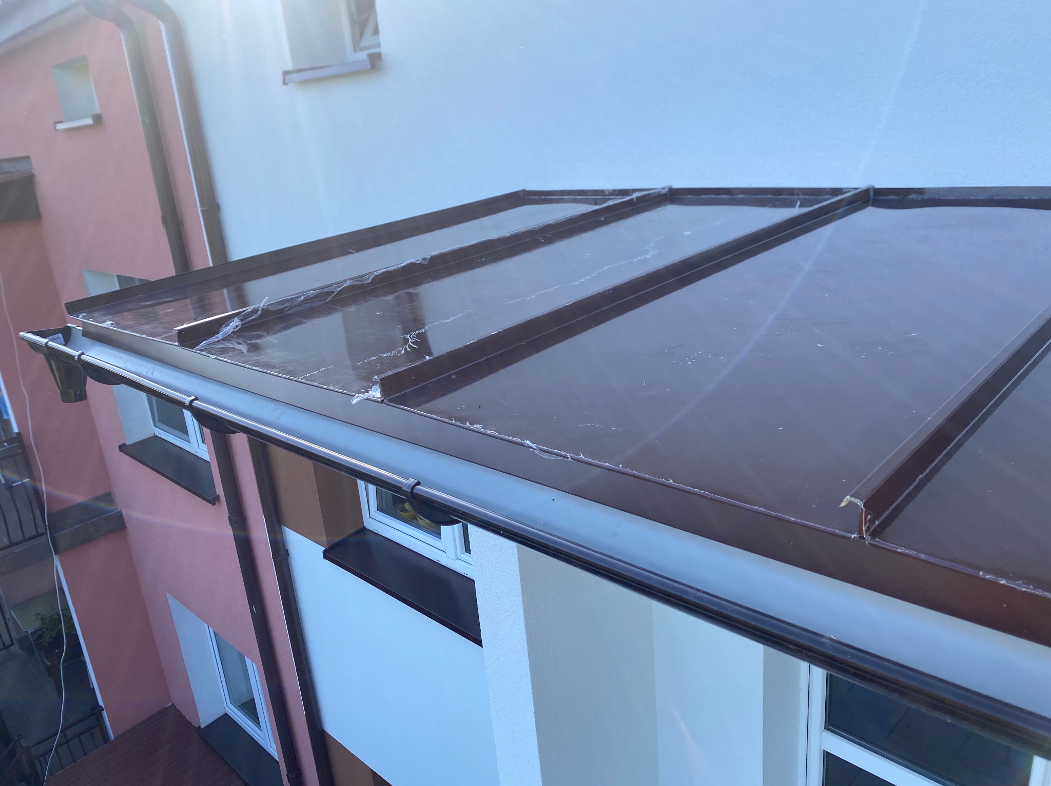 Błyszczące, brązowe pokrycie dachowe z rynnami na tle elewacji budynku w pastelowych kolorach. Widoczne pozostałości folii ochronnej na panelach.