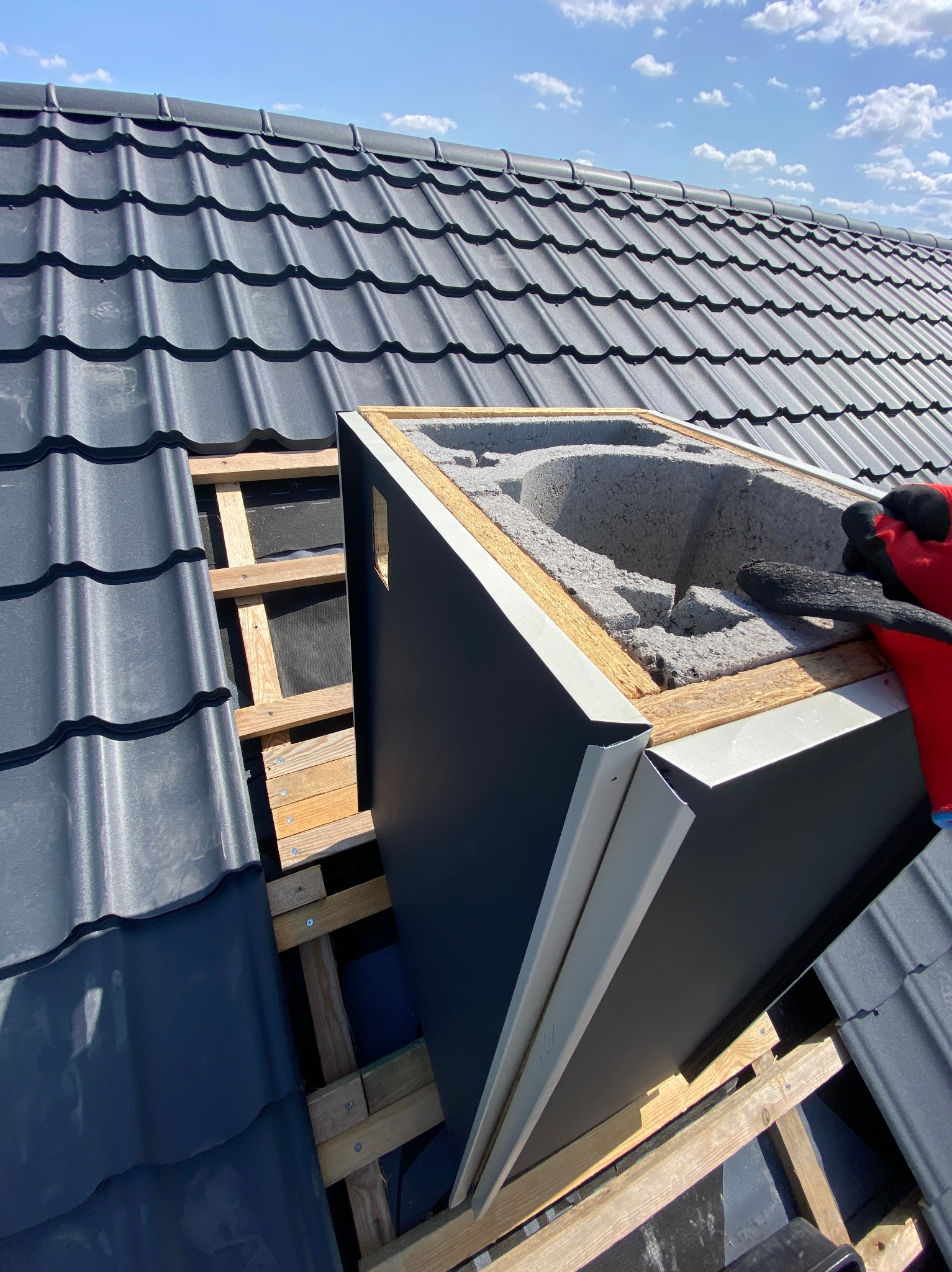 Montaż obróbki komina na dachu krytym szarą dachówką. Widoczna więźba i ocieplenie. Praca dekarza w trakcie, rękawice ochronne. Słoneczny dzień, błękitne niebo.