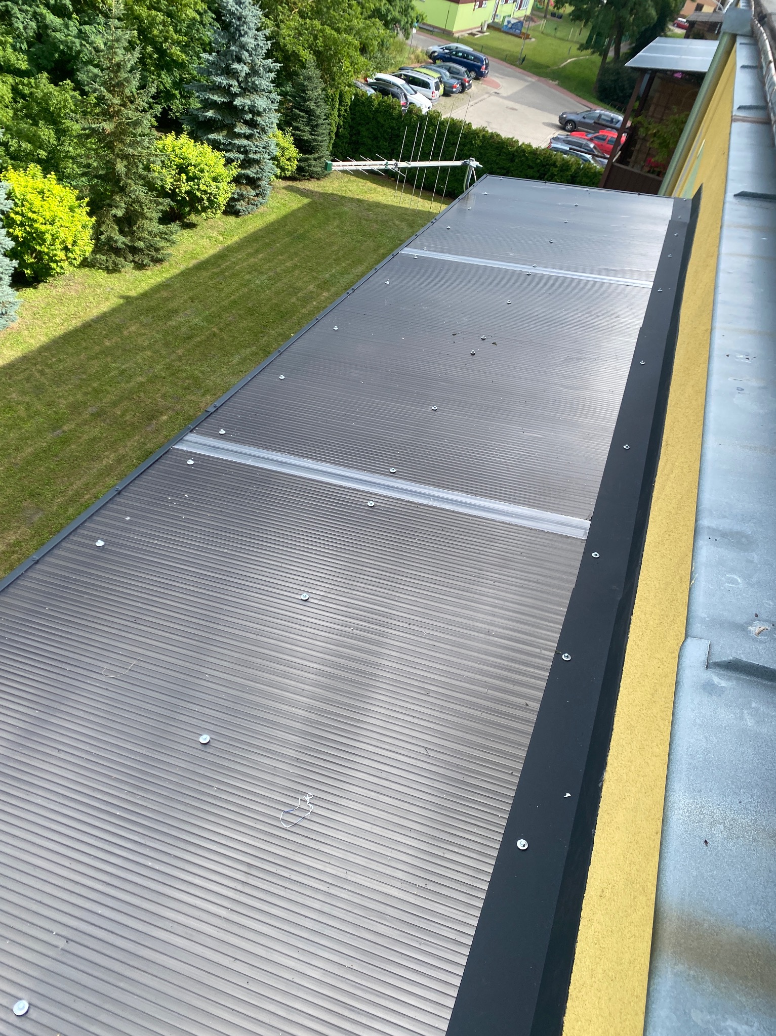 Szary dach z blachy trapezowej, zabezpieczony czarną listwą i przymocowany śrubami. Widok z góry na budynek i zieleń.