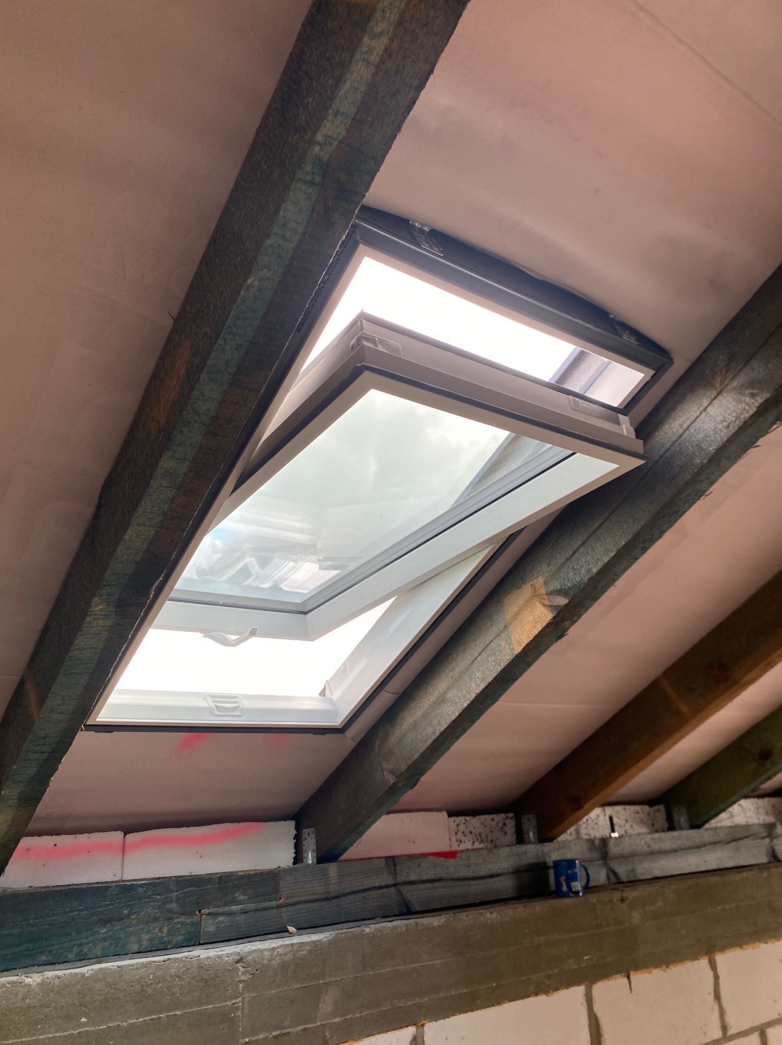 Otwarty, biały okno dachowe zamontowane w skośnym dachu z widoczną konstrukcją drewnianą i izolacją. Widok od wewnątrz, w tle jasne niebo.