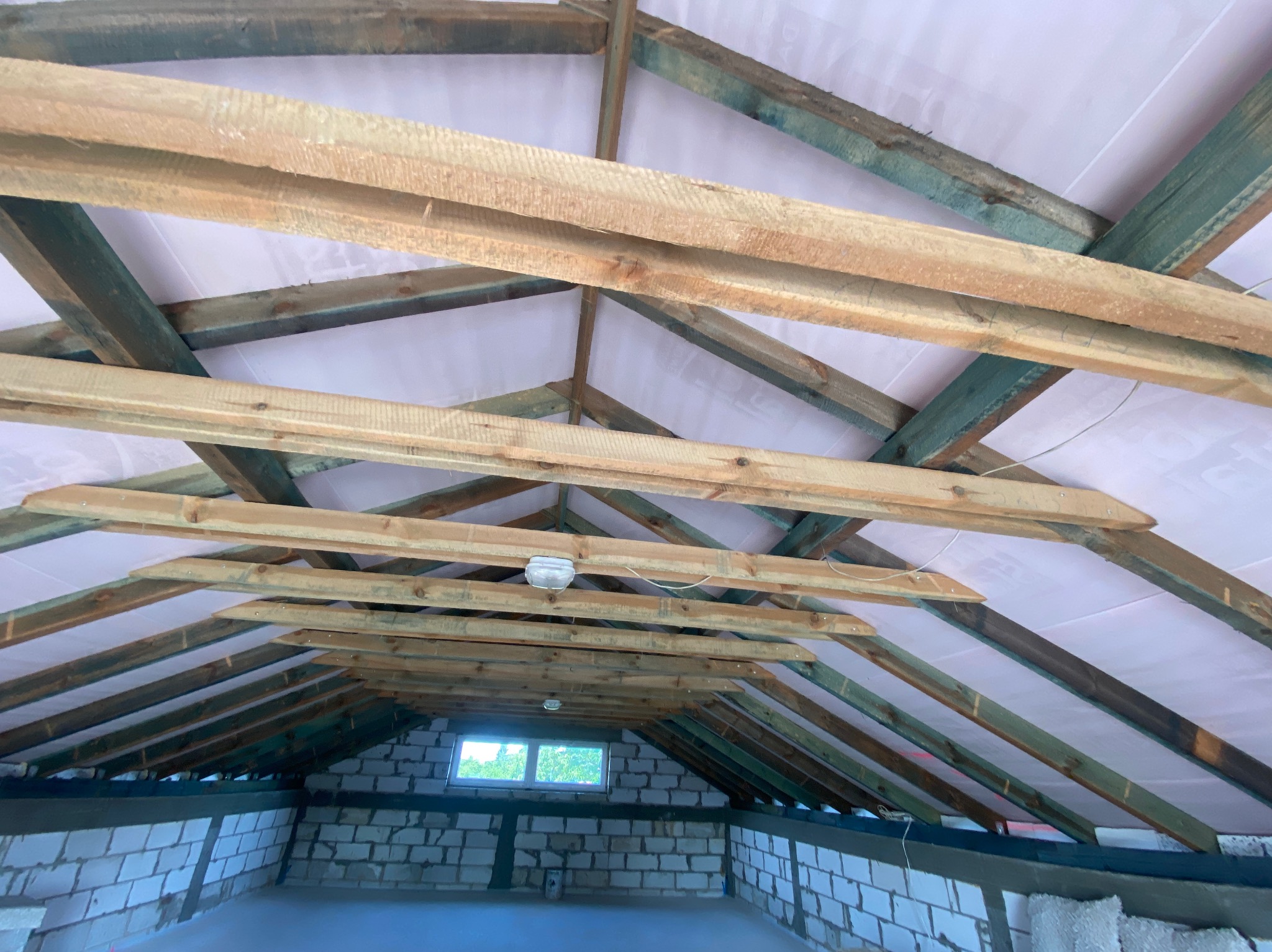 Wnętrze poddasza z widoczną drewnianą konstrukcją więźby dachowej i membraną. W oddali okno doświetlające przestrzeń. Widok z dołu.