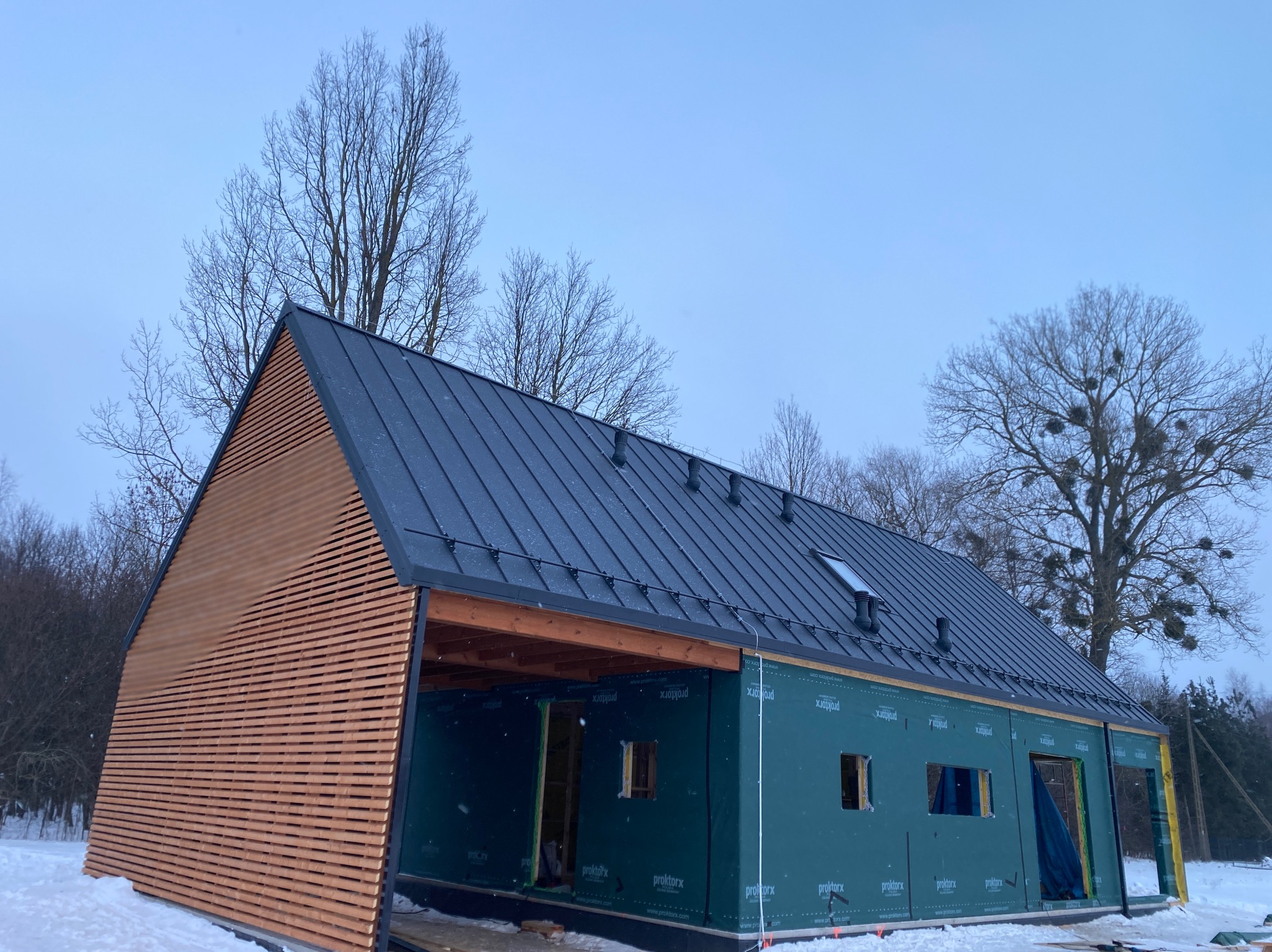 Nowoczesny dom w budowie z czarnym dachem i drewnianą elewacją. Widoczne membrany na ścianach i fragment zimowego krajobrazu w tle.