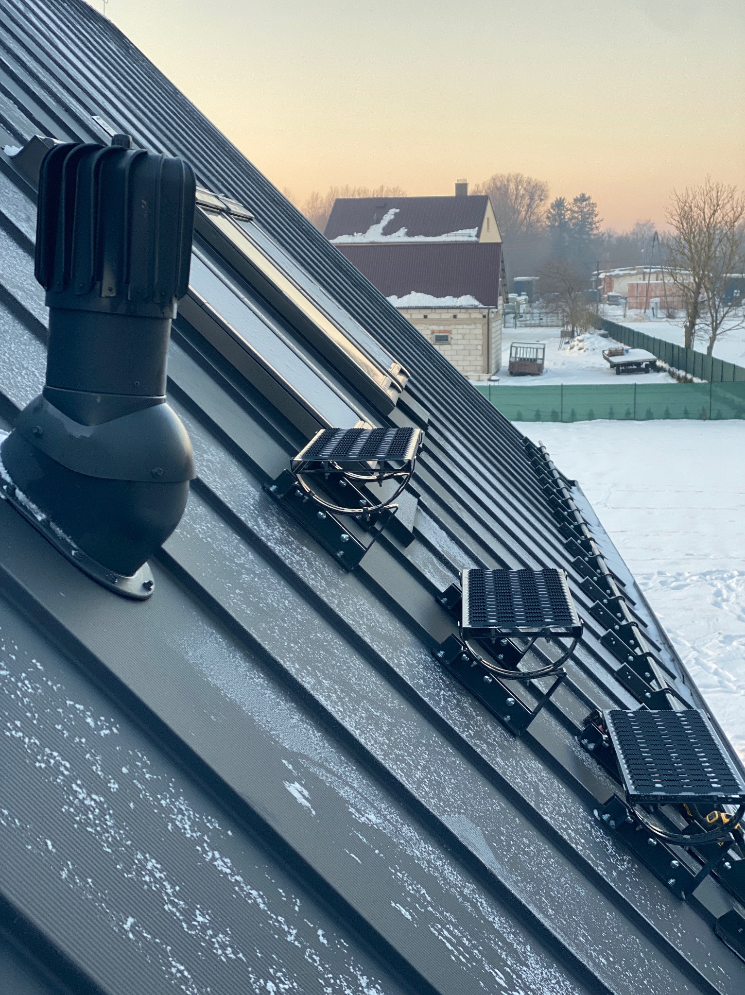 Szary dach z blachy trapezowej z zamontowanymi stopniami kominiarskimi i odpowietrznikiem. W tle ośnieżony krajobraz i budynki.