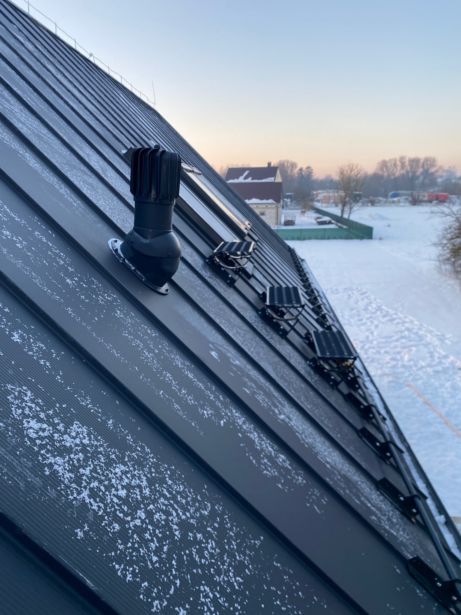 Czarny, metalowy dach pokryty śniegiem, z kominkiem wentylacyjnym i panelami solarnymi, na tle zimowego krajobrazu o zmierzchu. Widok z góry.