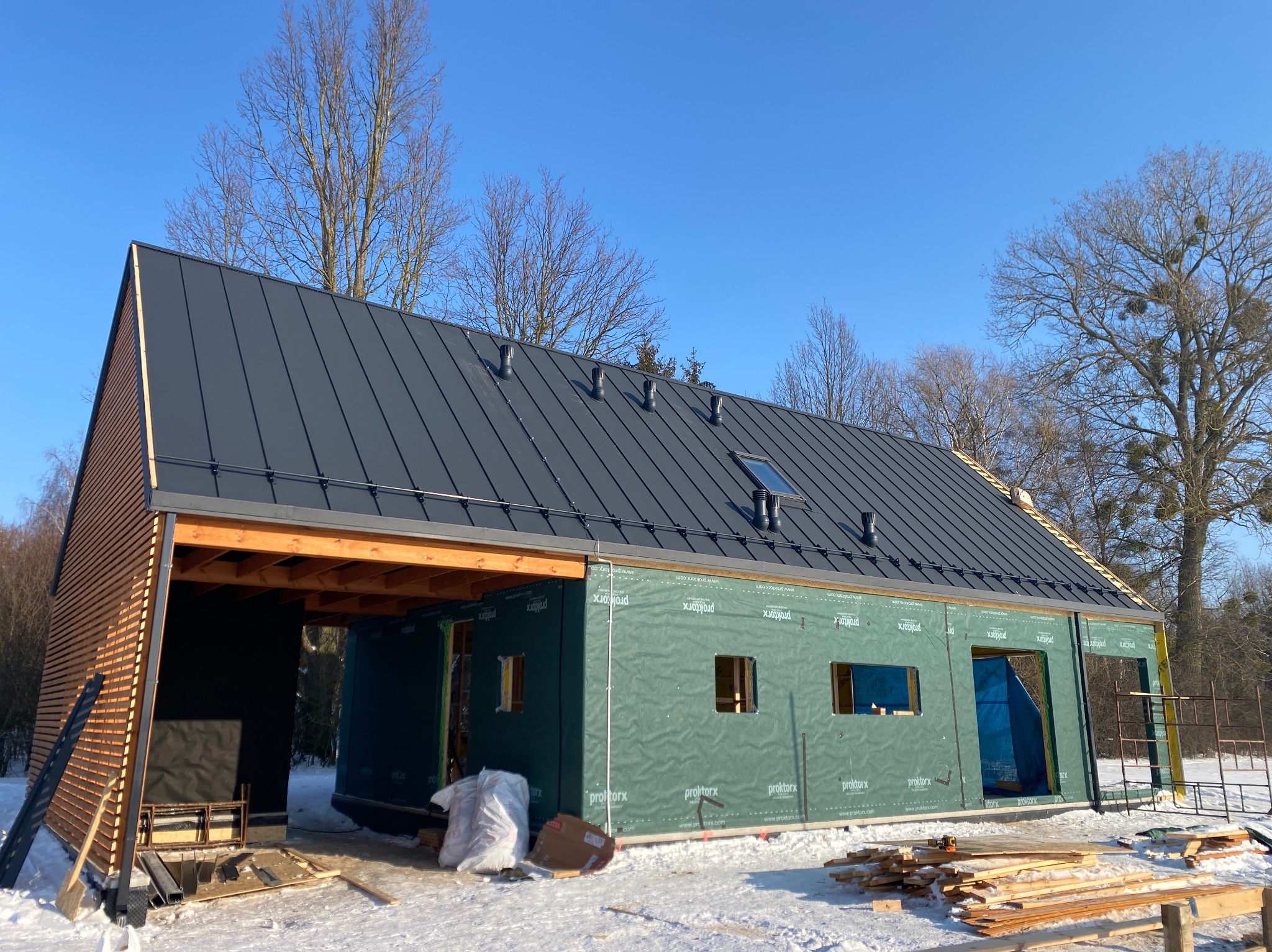 Nowoczesny dom w trakcie budowy, z czarnym dachem z blachy na rąbek stojący i drewnianą elewacją. Widoczne membrany na ścianach i elementy konstrukcyjne.