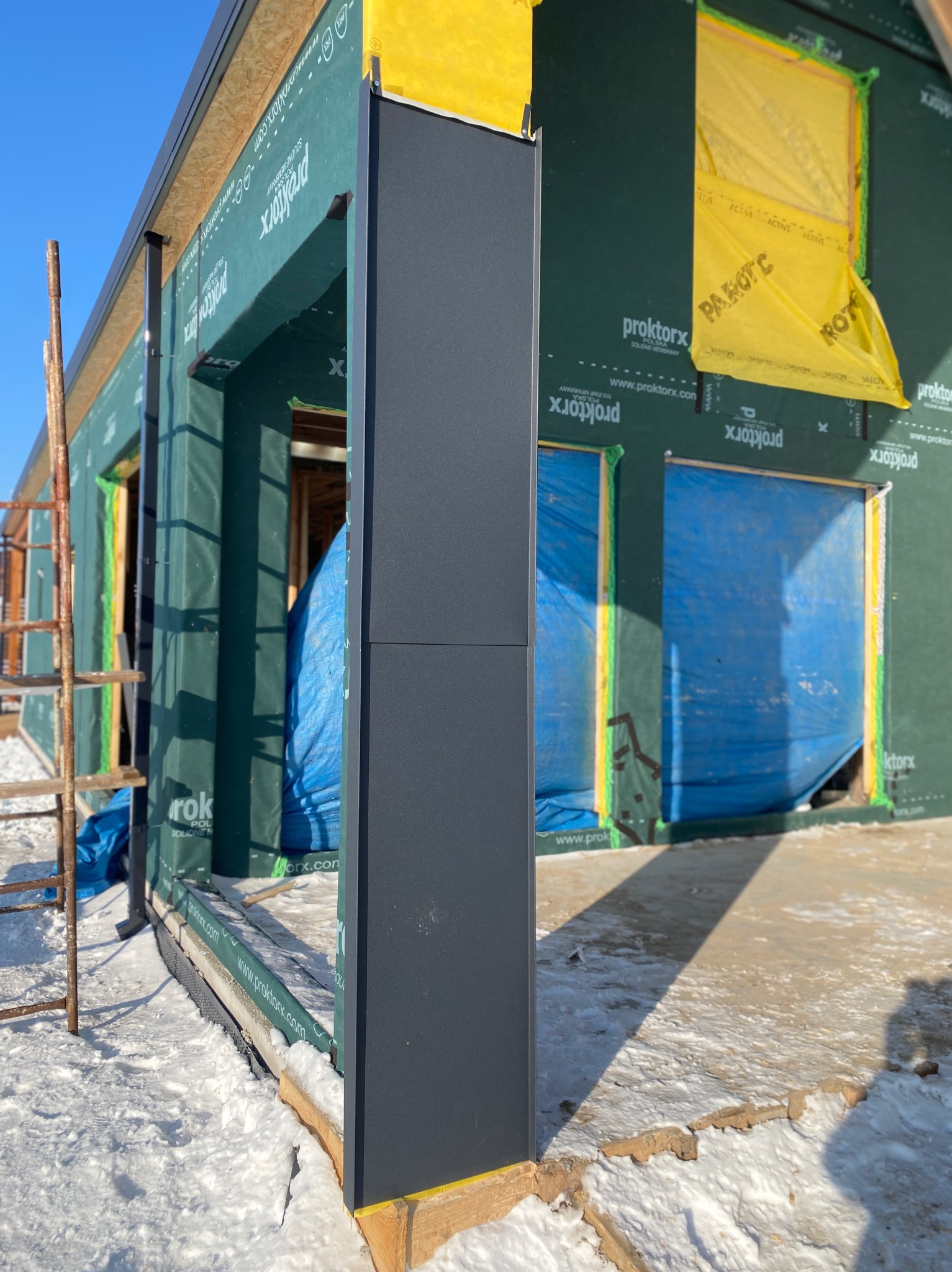 Budowa domu w trakcie: widoczna ściana z membraną, okna zabezpieczone folią, fragment rusztowania i ośnieżony teren. Elementy konstrukcyjne w kolorze grafitowym.