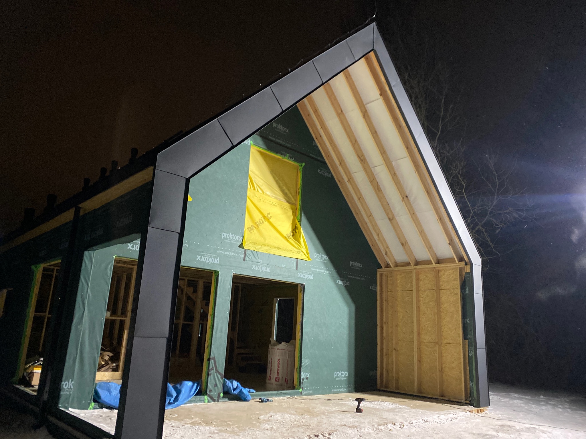 Budowa domu jednorodzinnego w Warszawie w trakcie prac dekarskich. Widoczna konstrukcja dachu z elementami izolacji i folii. Ujęcie nocne, oświetlenie budowlane.