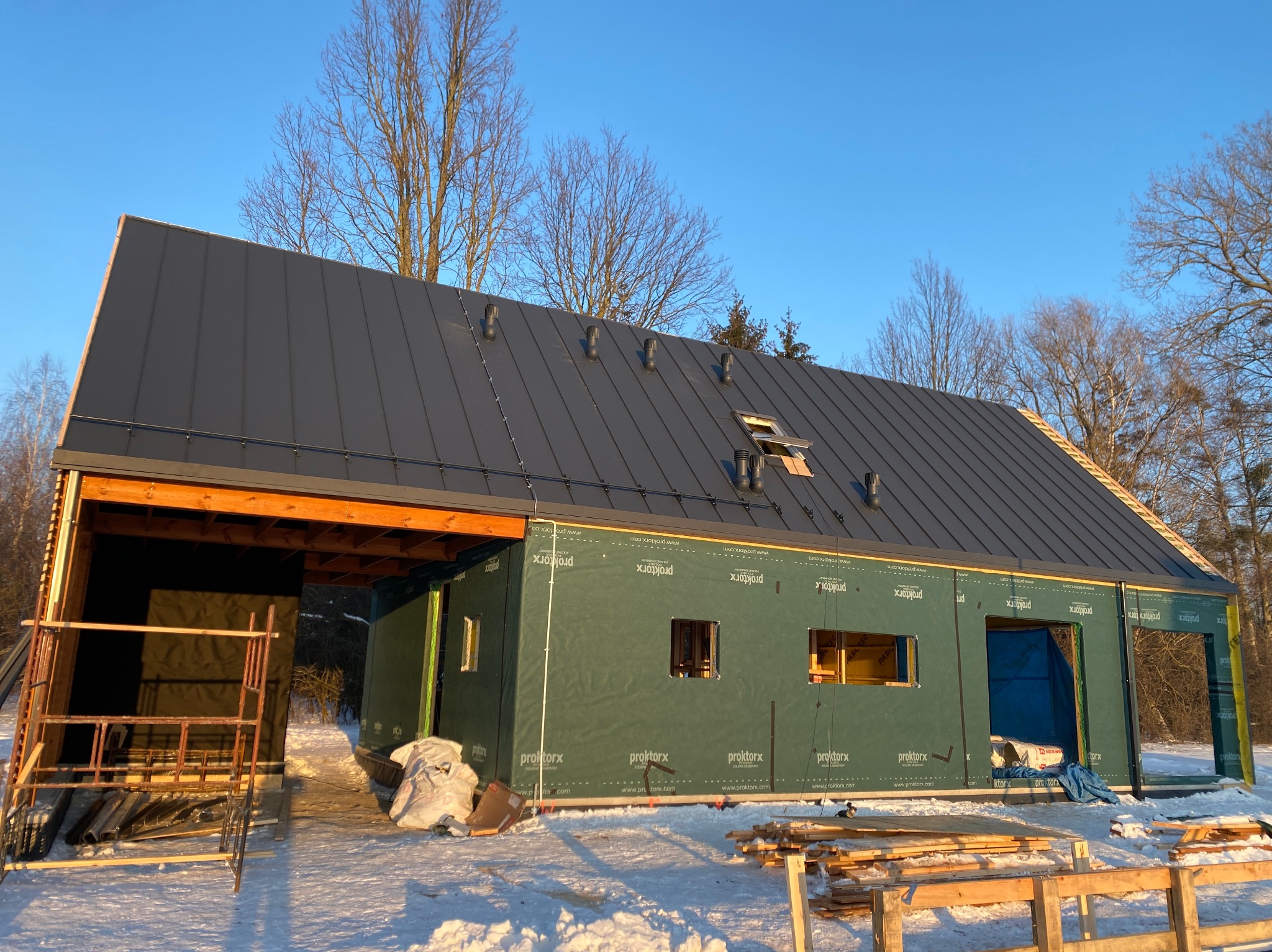 Budowa domu w zimowej scenerii: szary dach z blachy na budynku w stanie surowym zamkniętym, widoczne rusztowania i materiały budowlane, błękitne niebo w tle.
