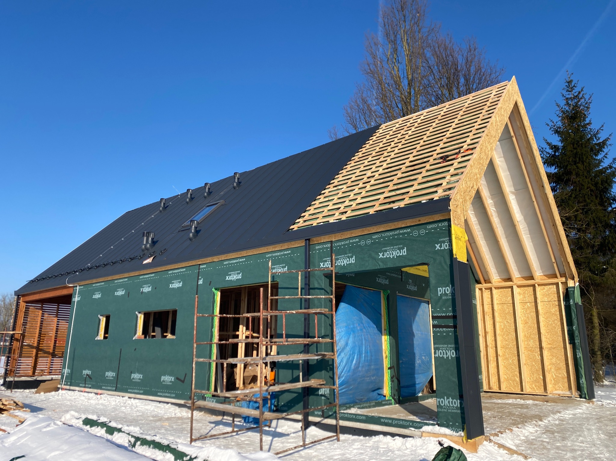 Budowa dachu domu jednorodzinnego w zimowej scenerii. Część dachu pokryta blachą, druga część z widoczną konstrukcją drewnianą. Widoczne okna dachowe i membrany.