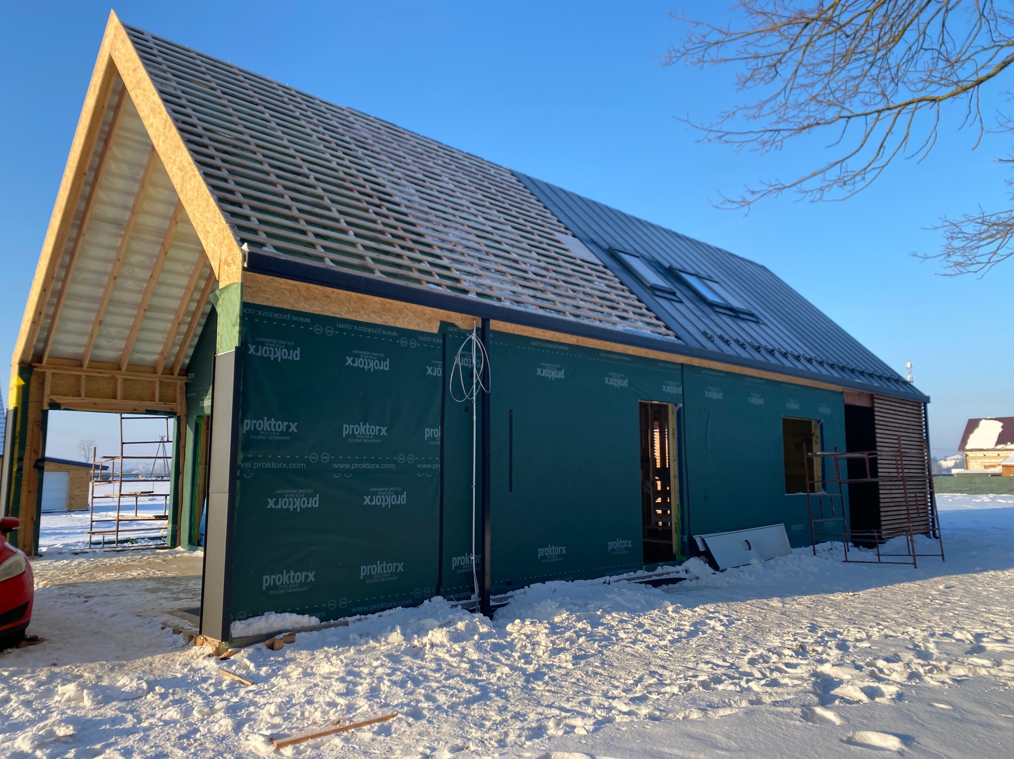 Budowa domu w zimowej scenerii. Częściowo pokryty dach z widoczną więźbą. Zabezpieczona ściana folią. Widoczne rusztowania i śnieg.