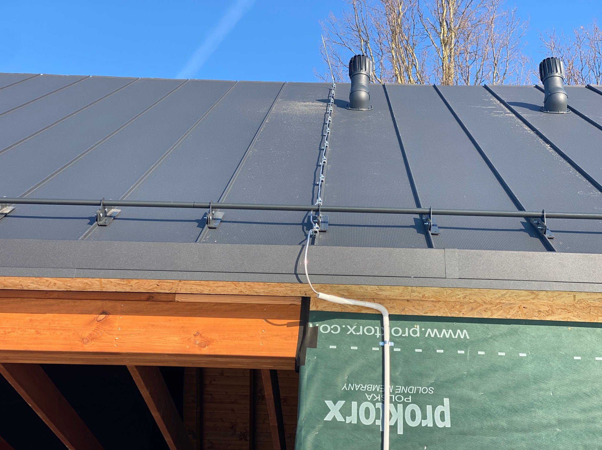 Szary dach z blachy na rąbek stojący, z systemem odgromowym i kominkami wentylacyjnymi. Widoczne elementy konstrukcji dachu i membrana dachowa.