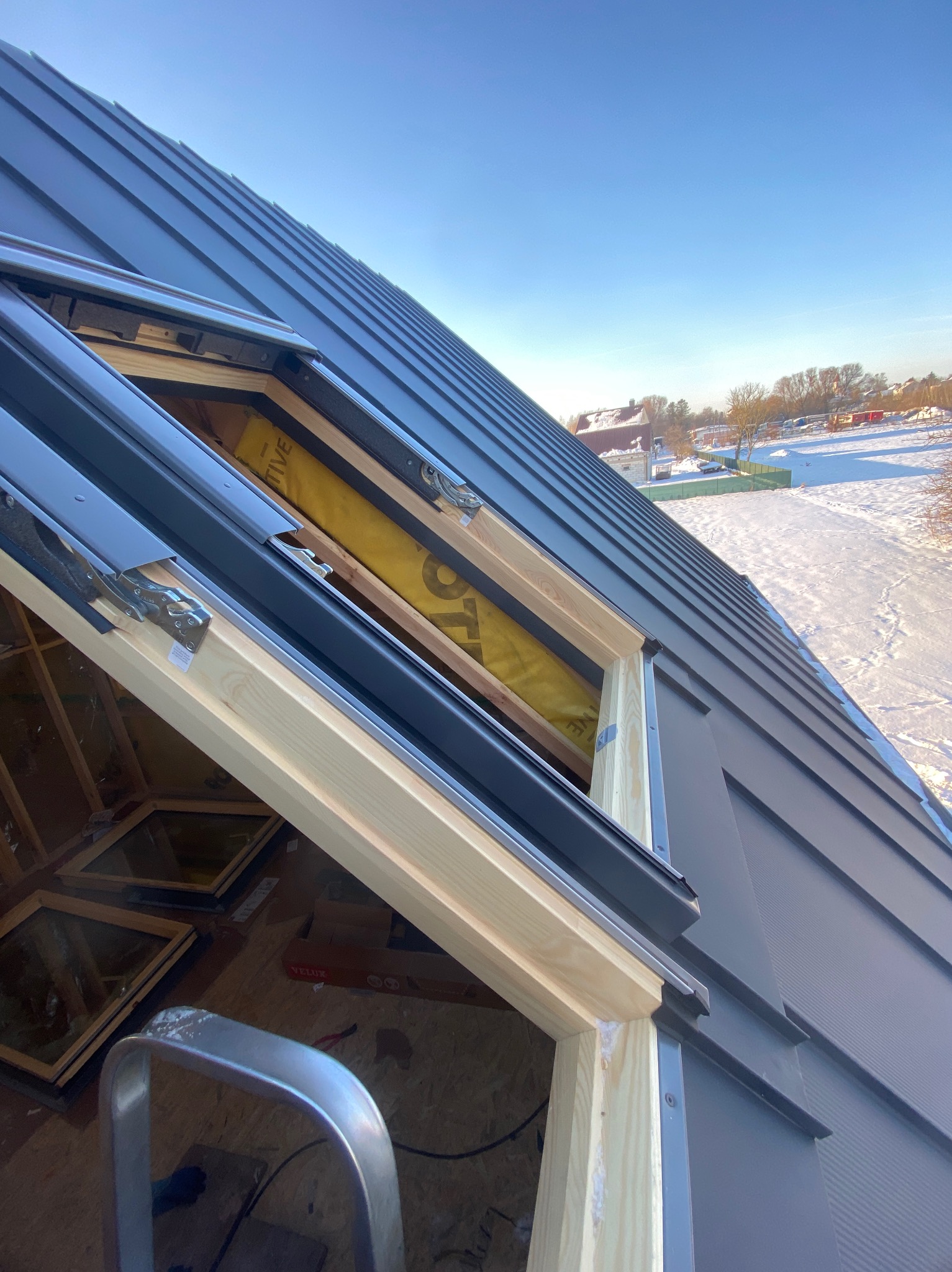 Montaż okna dachowego w pokryciu z blachy na tle zimowego krajobrazu. Widoczne drewniane ramy okienne i fragment drabiny.