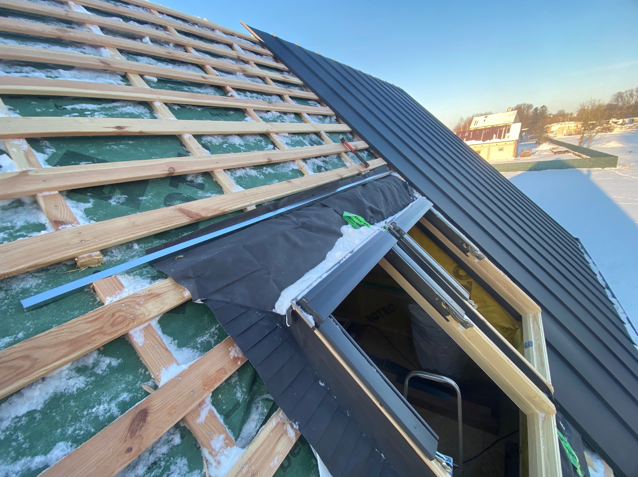 Montaż okna dachowego w pokryciu z blachy na drewnianej konstrukcji dachu, widoczne ośnieżenie i membrana dachowa. Częściowo ukończony etap prac dekarskich.