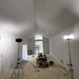 MydMar - Wnętrze w trakcie malowania natryskowego na biało. Widoczny agregat malarski, reflektory na trójnogach i zabezpieczenia okna. Ściany i sufit pokryte świeżą farbą.