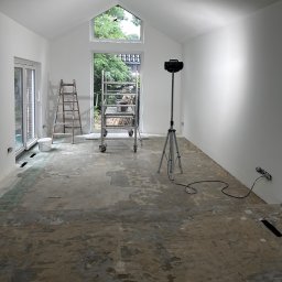 MydMar - Wnętrze w trakcie remontu: białe ściany, surowa podłoga, drabiny i oświetlenie budowlane. Widok na zieleń przez okno.