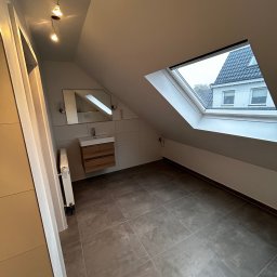 MydMar - Nowoczesna łazienka na poddaszu z oknem dachowym, szarymi płytkami, drewnianą szafką pod umywalką i minimalistycznym oświetleniem. Widok na budynek przez okno.