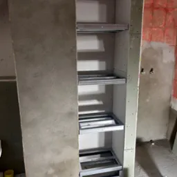 Szkielet regału z płyty GK z metalowymi profilami na półki, widoczne narzędzia i poziomica. Ściana w tle przygotowana pod płytki.