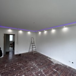 MydMar - Białe wnętrze z zamontowanym oświetleniem LED przy suficie, drabina w rogu pokoju, ślady farby na podłodze, otwarte drzwi do kolejnego pomieszczenia.