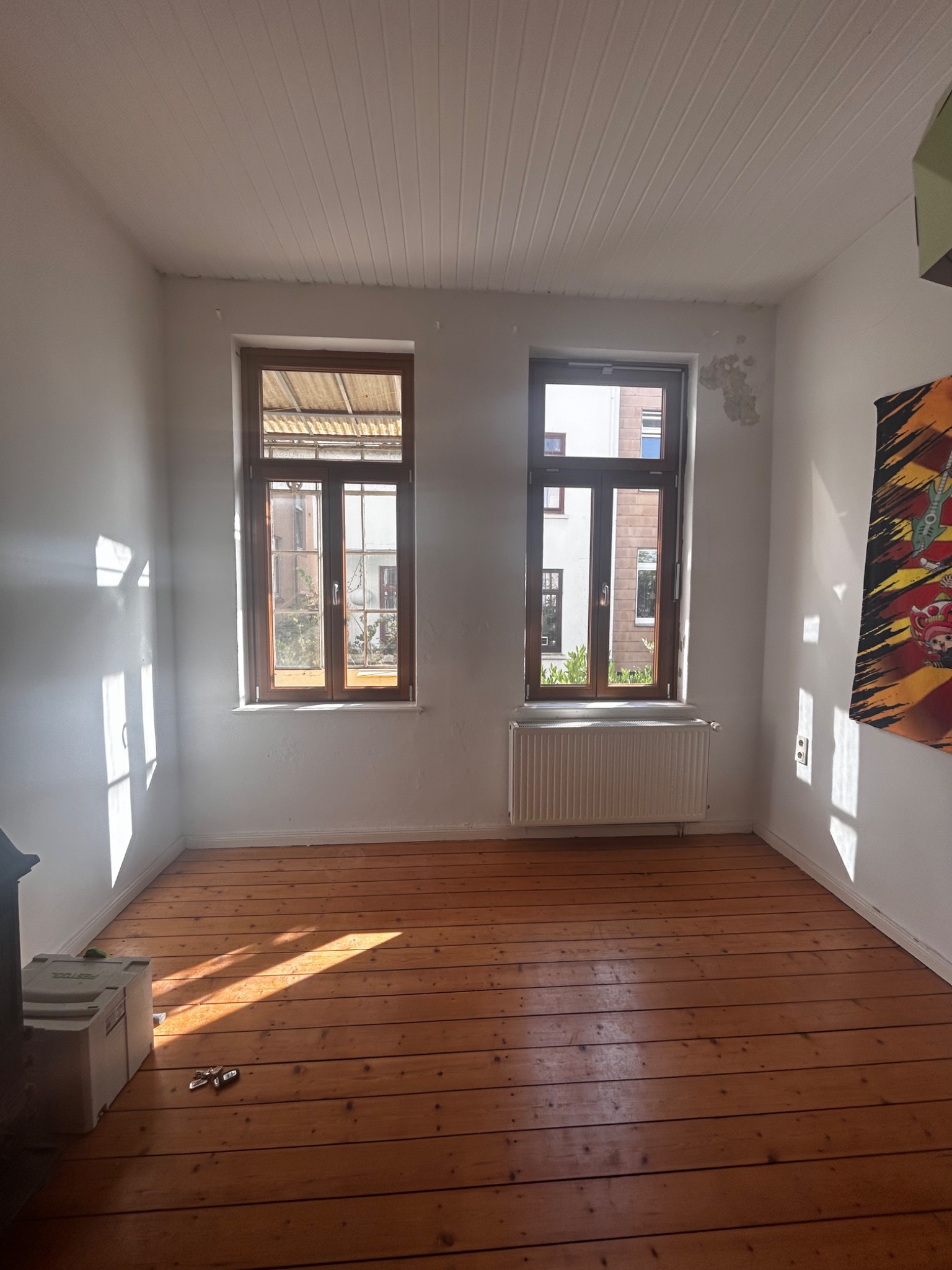 Pusty pokój z drewnianą podłogą i dwoma oknami, widać grzejnik. Na ścianie ślady po usuniętej tapecie, a na podłodze narzędzia i skrzynka narzędziowa.