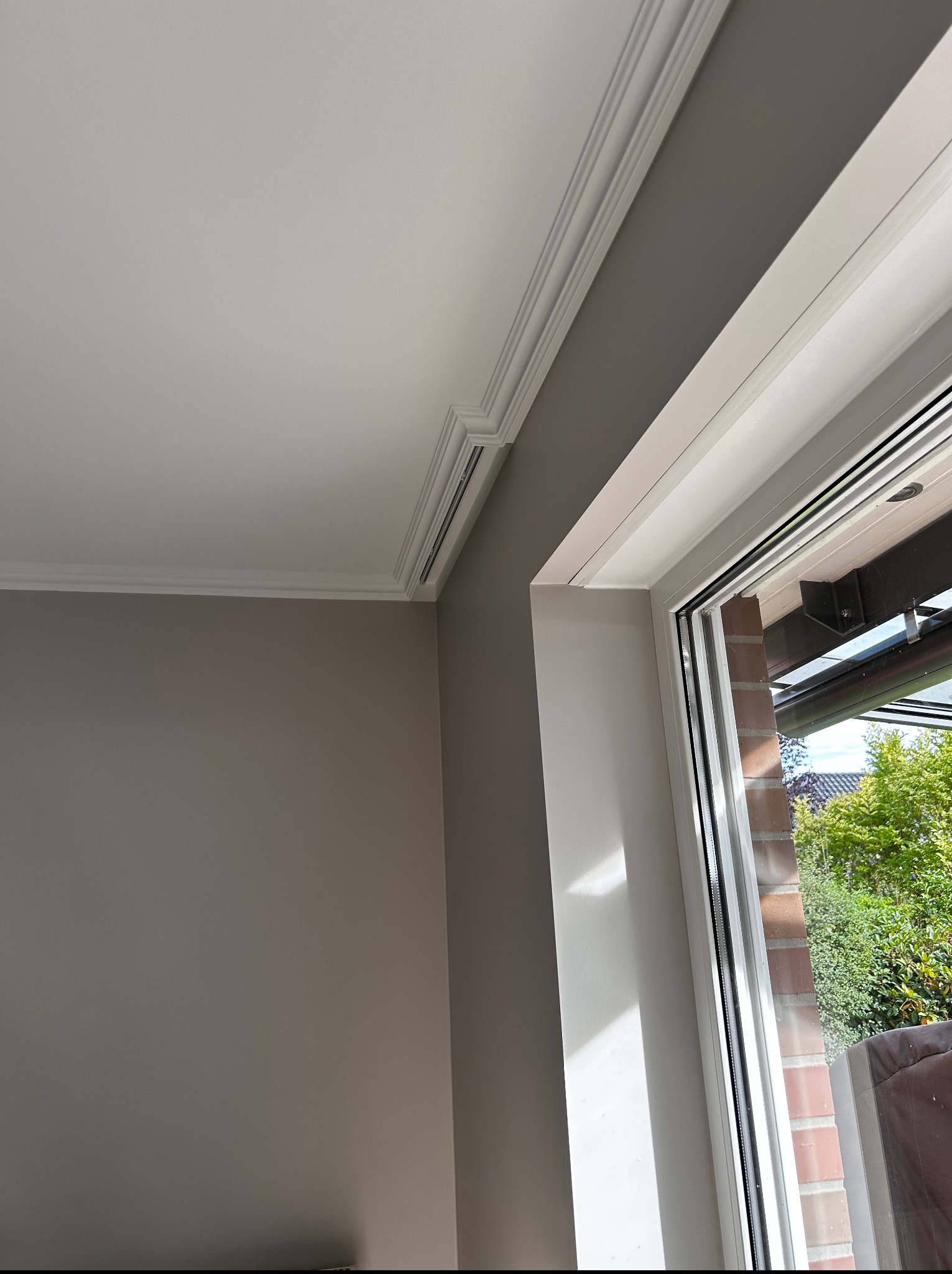Wykończony pokój z oknem: szare ściany, biały sufit z ozdobną listwą i biała rama okienna z widokiem na zieleń. Detal wykończenia wnętrza.
