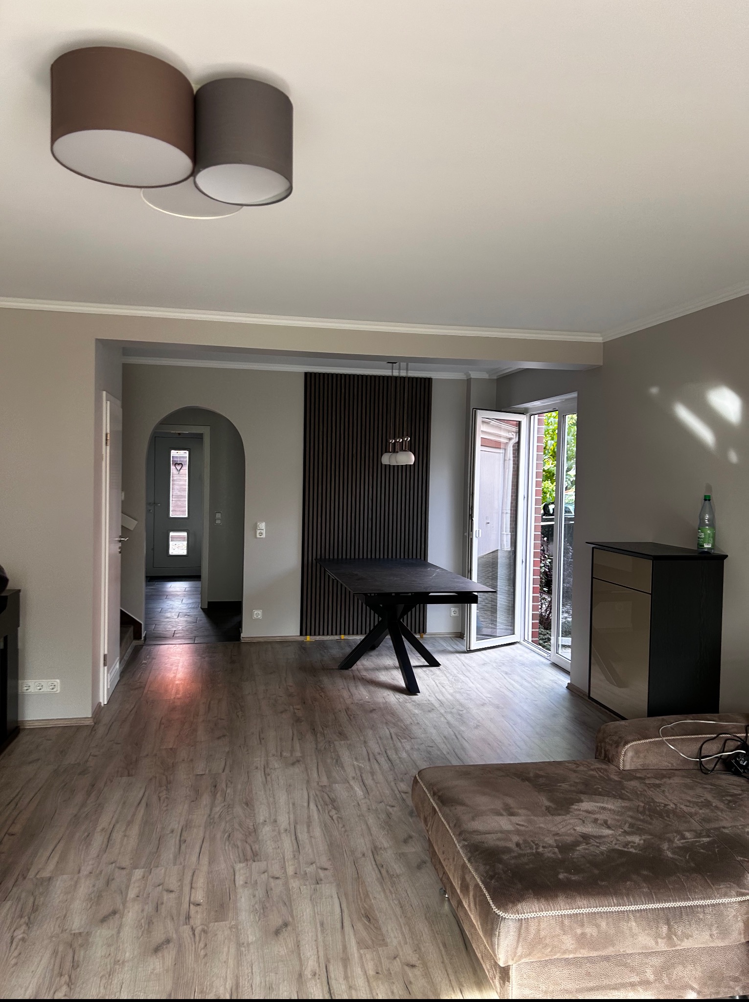 Jasne wnętrze z drewnianą podłogą, ciemnym stołem i dekoracyjną ścianą z pionowych paneli. Widoczne drzwi balkonowe i oświetlenie sufitowe.