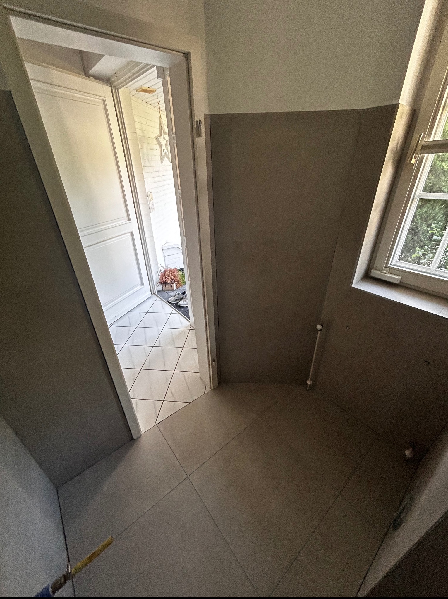 Wykończony przedpokój z szarą ścianą, jasnymi płytkami podłogowymi i otwartymi drzwiami wejściowymi. Widoczne okno z widokiem na zieleń.