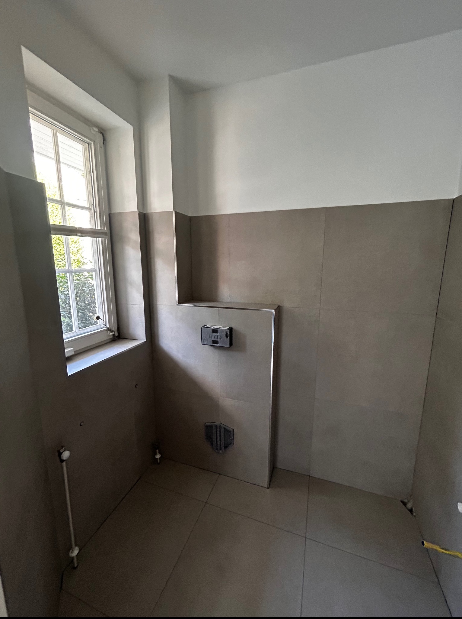 Wykończona łazienka z szarymi płytkami, widoczny stelaż podtynkowy WC i okno z białymi ramami. Minimalistyczny design, surowe wykończenie.